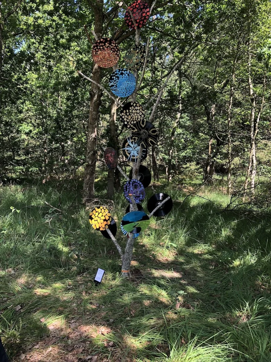 Kunstinstallation i form af en træ med farverige keramiske skiver med mønstre i skovmiljø.