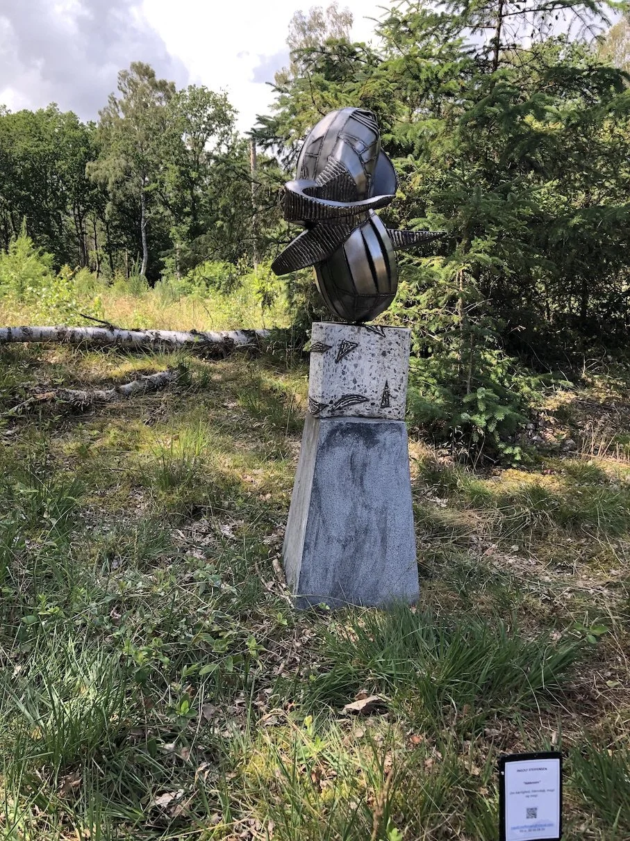 Skulptur af en sort, modern stil, formet som en blomst eller en frugt, placeret på en grå sten-tromle i en skov med grønne træer og græs.