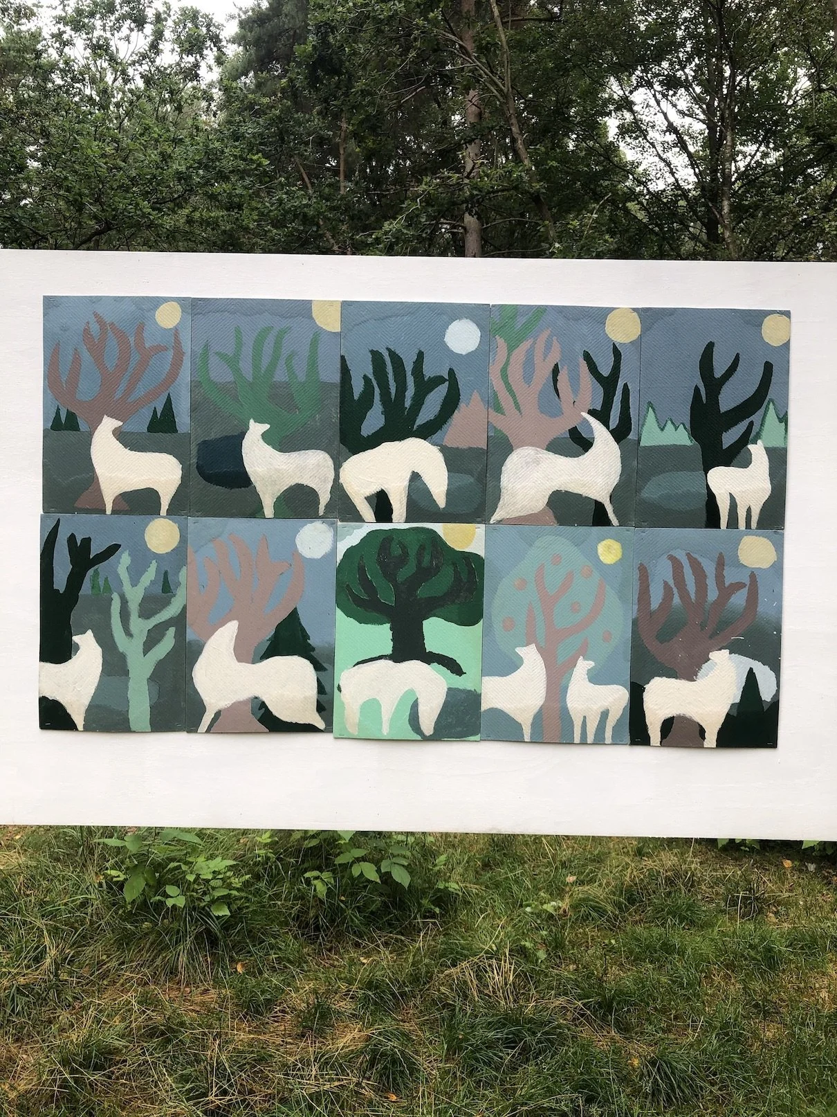 En collage af tolv malerier med ugler, træer og måner, alle i pastel- og jordfarver, sat op på en hvid ramme udendørs.
