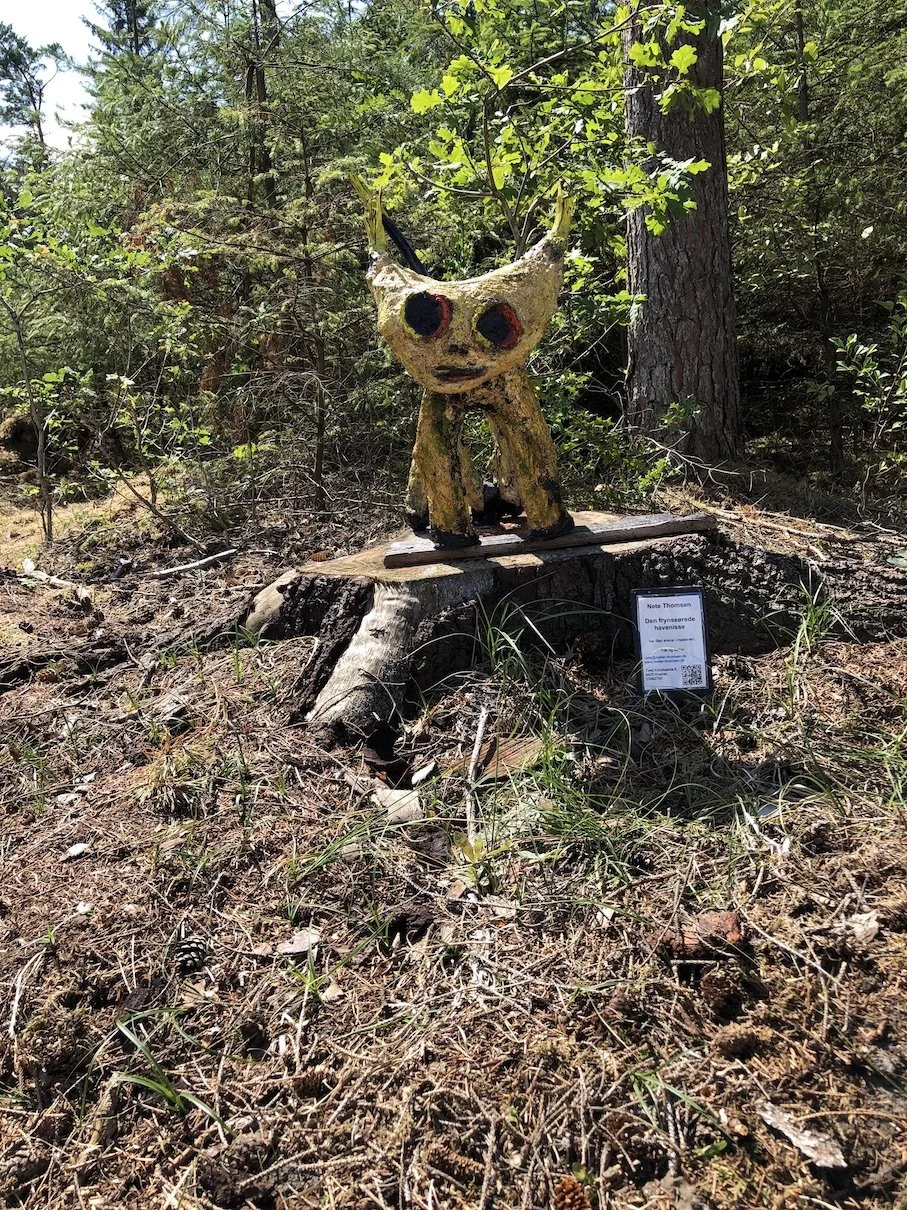 En skulptur af en fantasifigur, der ligner en kat med store øjne og lys hudfarve, placeret på en træstub i en skov.