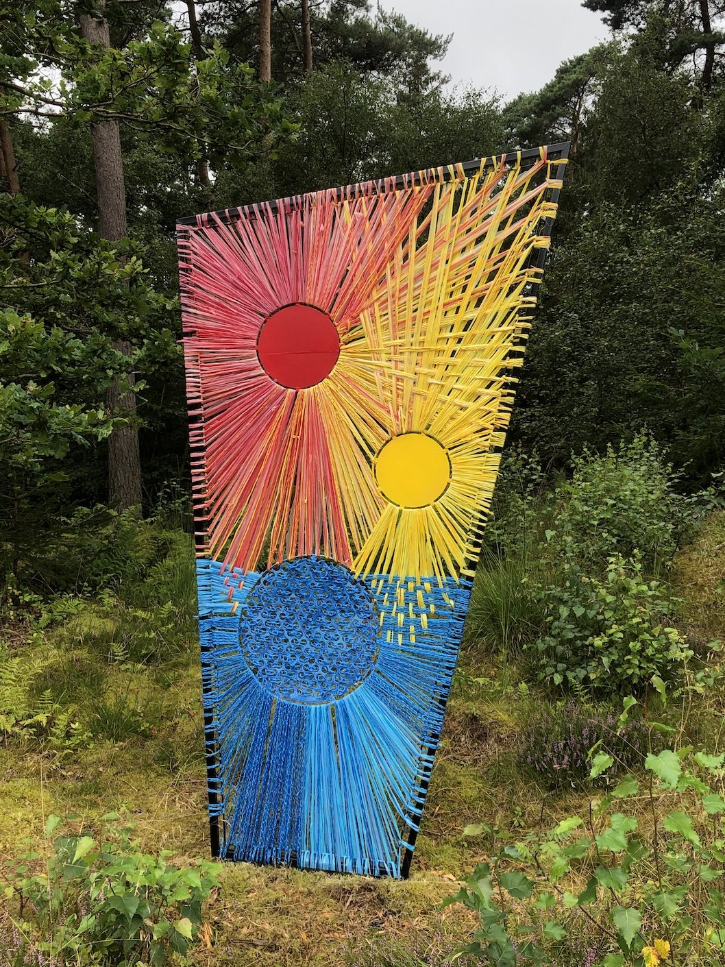 Farvet kunstværk med sol og måne motiv, lavet tråde, placeret i en grøn skov.