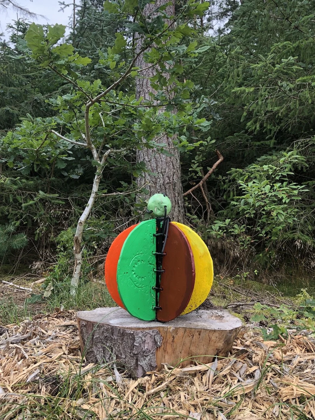 Farvet, kunstfærdig skulptur med grønt, brunt, gult og orange farver, placeret på en træstub i en skov.
