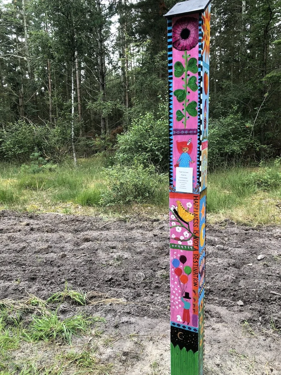 Farverig, ligner en kunstinstallation eller vejskilt med forskellige illustrationer, herunder en blomst, firkløver, en pige med hat og balloner, omgivet af træer i en skov.