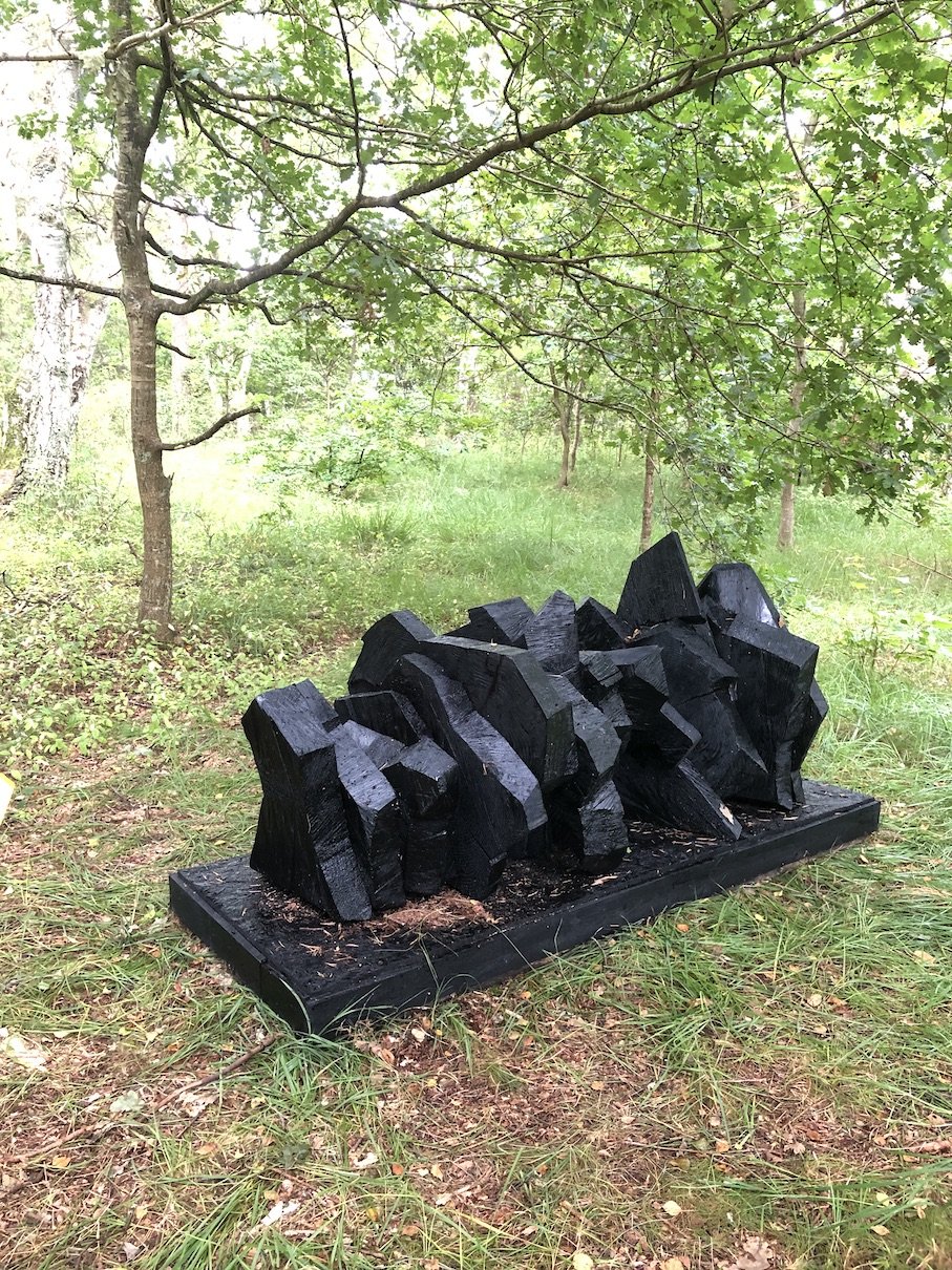 Sort træblokke er stablet på en sort platform i en skov med grønne træer og græs omkring.