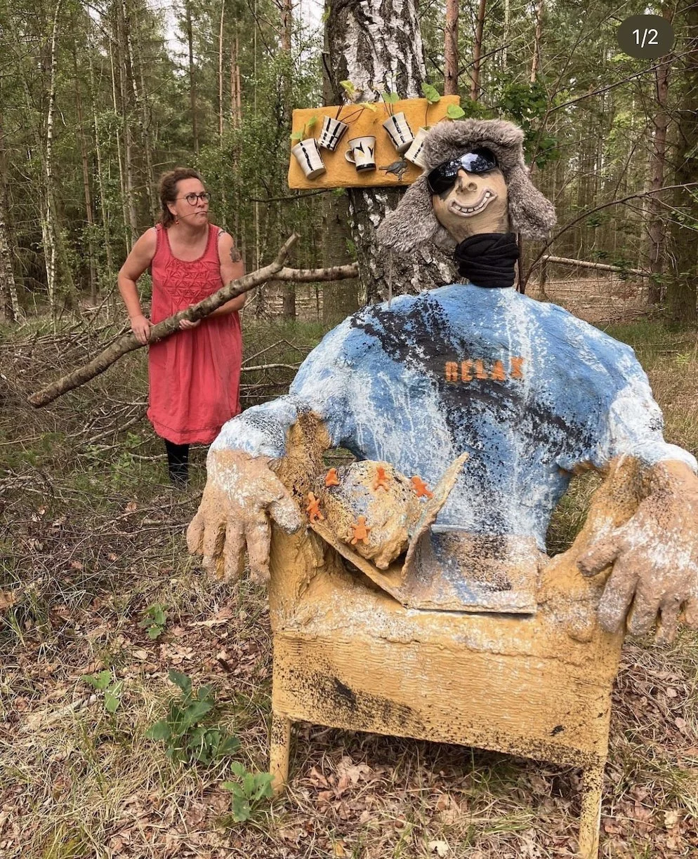 En kvinde i en rød kjole står i en skov ved siden af en stor kunstfærdig figur, der forestiller en person med solbriller, iklædt en blå t-shirt og har en stor grinende mund.