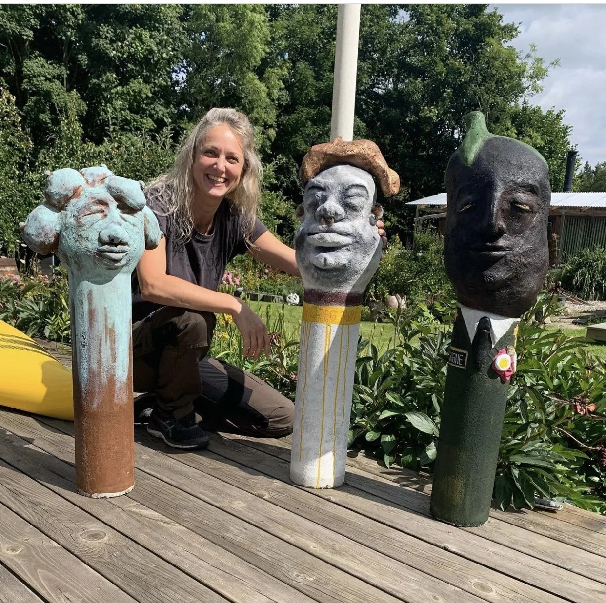 En smilende kvinde med blond hår, iført sort t-shirt og bukser, sidder på en træterrasse med tre kunstneriske skulpturer med hoveder, omgivet af grønt område med træer og planter.