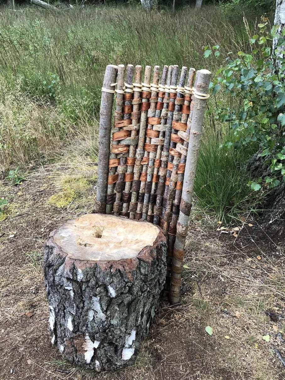 En stol lavet af en træstamme og en ryglæn lavet af små stykker træ og grene bundet sammen med reb, placeret i en skov.