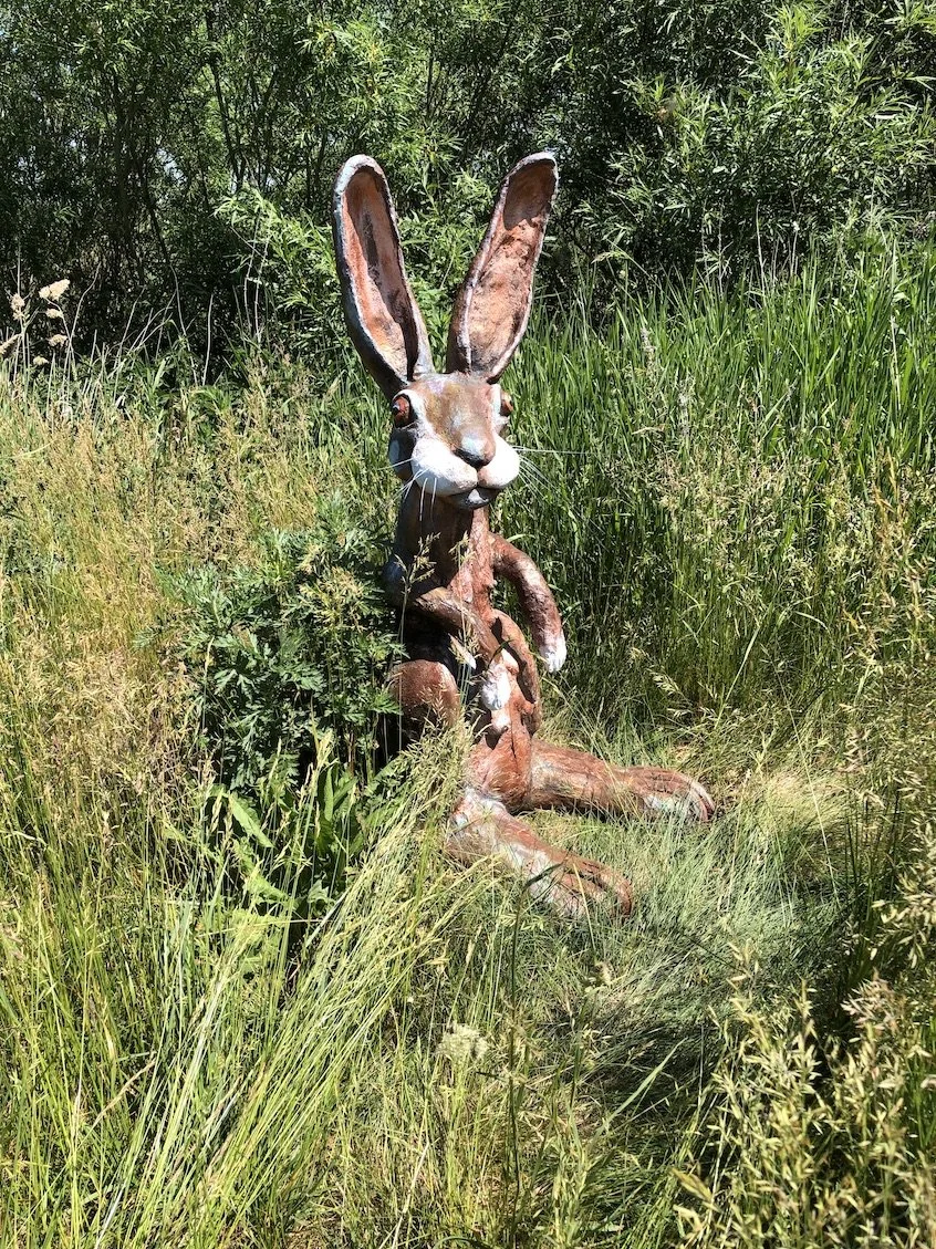 En stor hare-skulptur placeret i naturen blandt højt græs og buske.