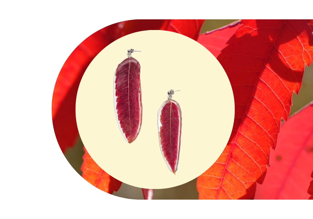 Red leaf-shaped earrings with silver borders and leaf vein details, displayed on a yellow background with red autumn leaves in the background.