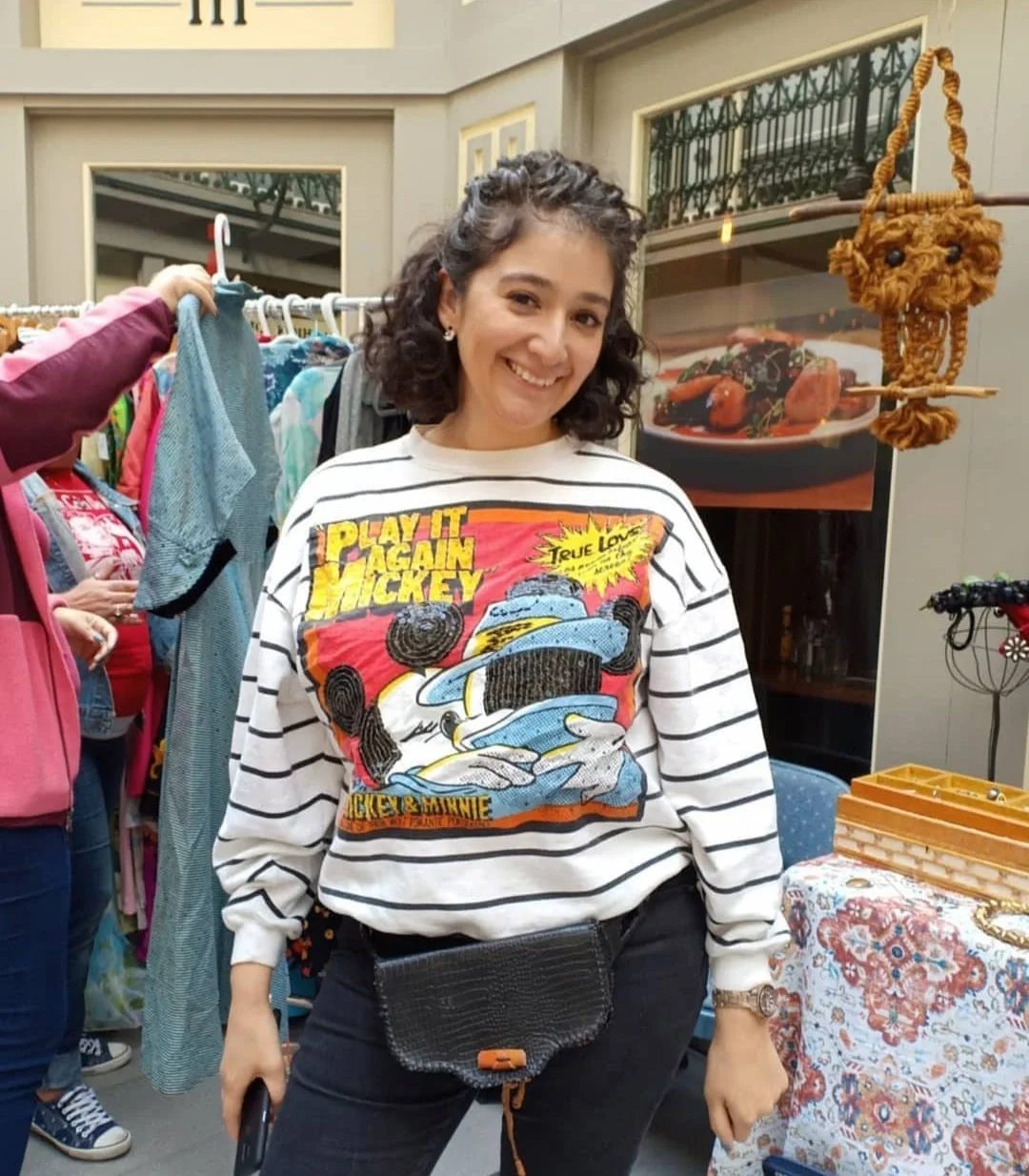 A woman smiling at a market or thrift store with clothing racks behind her and various decorative items nearby.