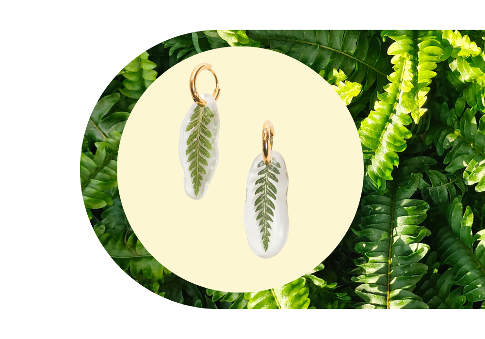 White oval earrings with green fern leaf design, set against a background of green fern leaves.