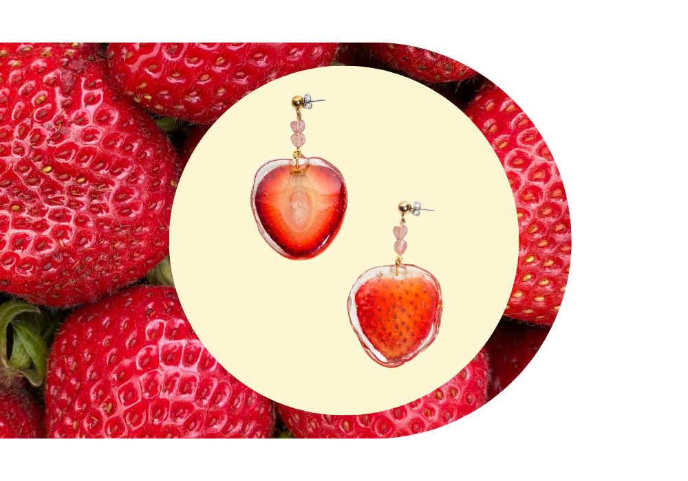 Strawberry-shaped earrings with pink heart beads on gold chains, set against a background of fresh strawberries.