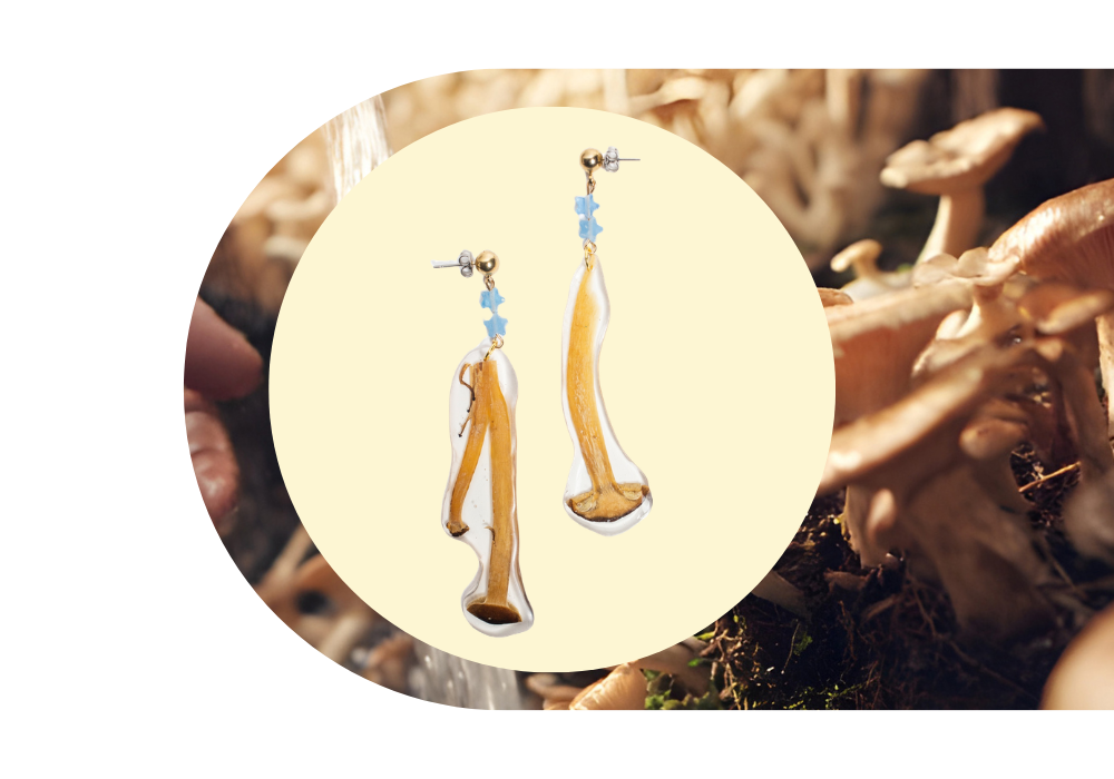 A pair of earrings with elongated, irregularly shaped pendants resembling dried mushroom stems, hanging from small blue bead clusters on gold and silver studs, placed against a yellow circular background over a backdrop of mushrooms.
