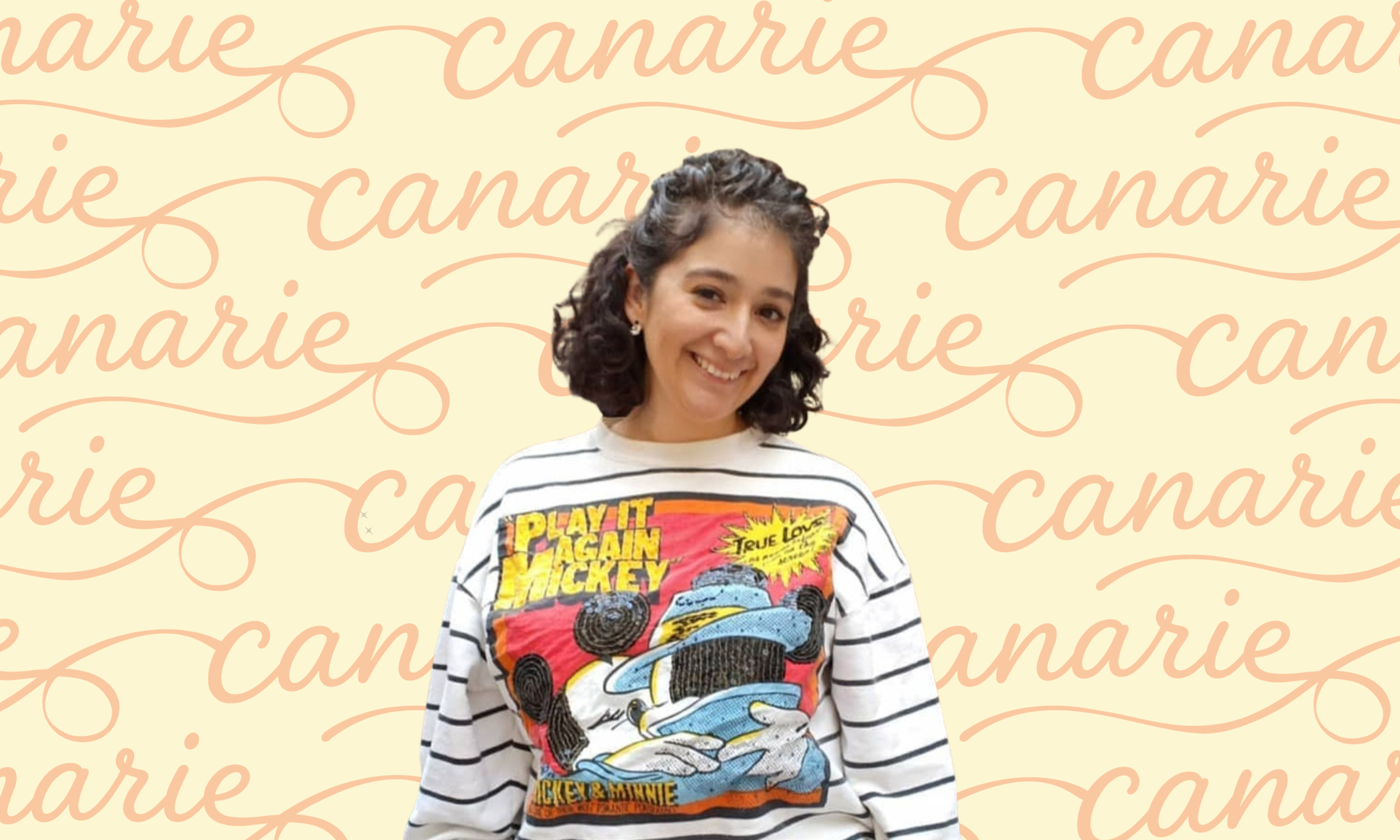 A young woman with curly dark hair smiling in front of a beige background with a repeating pattern of the words 'canarie' written in cursive pink font. She is wearing a white and black striped shirt with a colorful graphic featuring Mickey Mouse and Minnie Mouse on it.