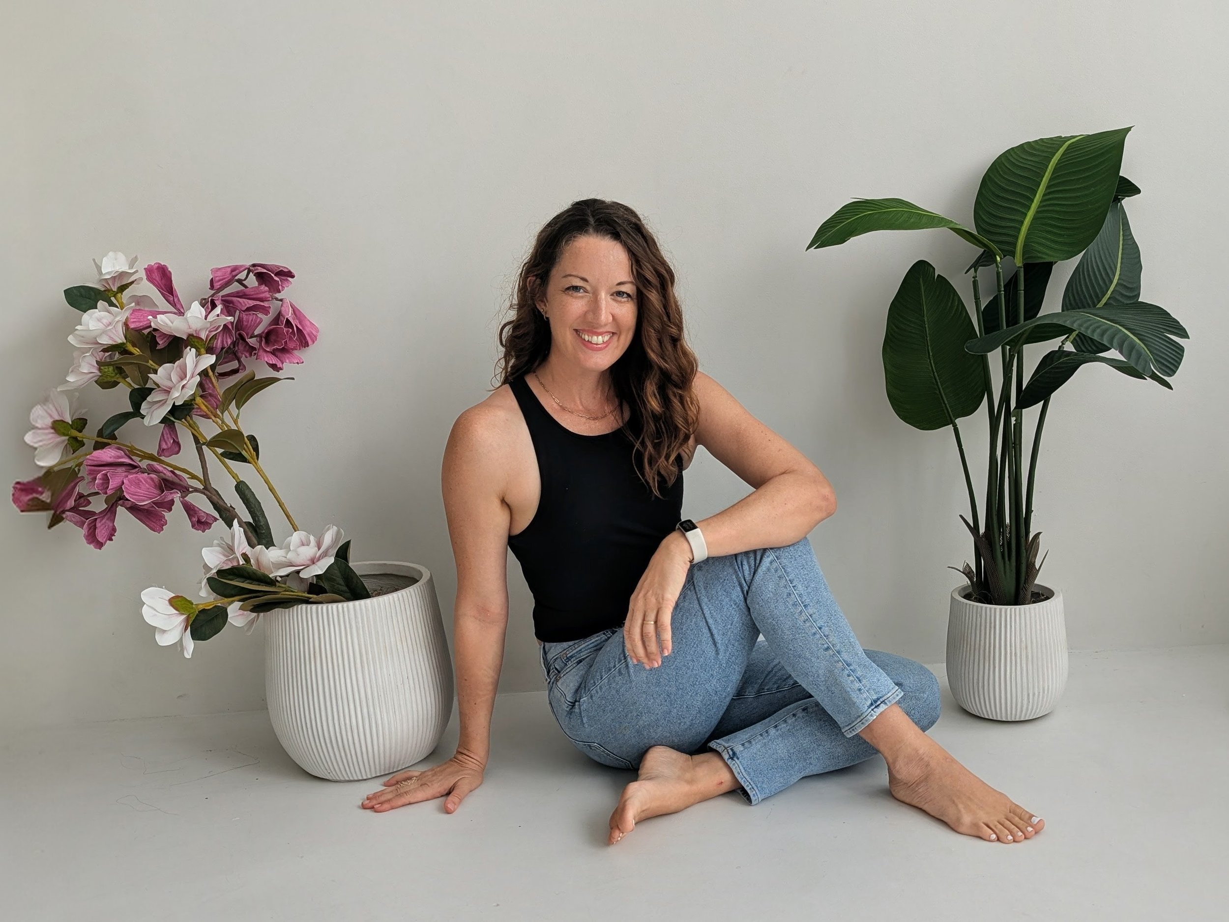 Woman with wavy brown hair sitting on the floor between two potted plants, wearing a black sleeveless top and blue jeans, smiling at the camera.