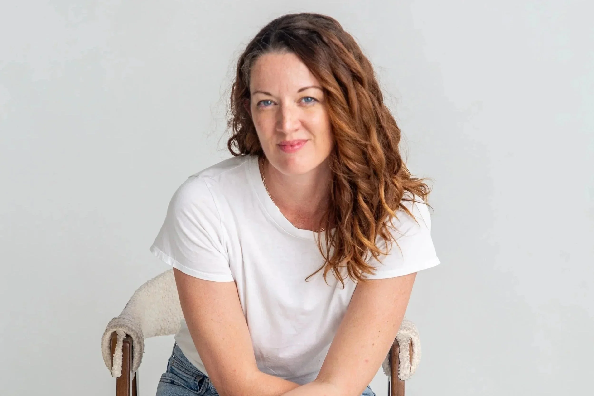 Joanie Johnson with long, curly, auburn hair and blue eyes, wearing a white t-shirt, sitting on a beige chair against a plain light gray background.