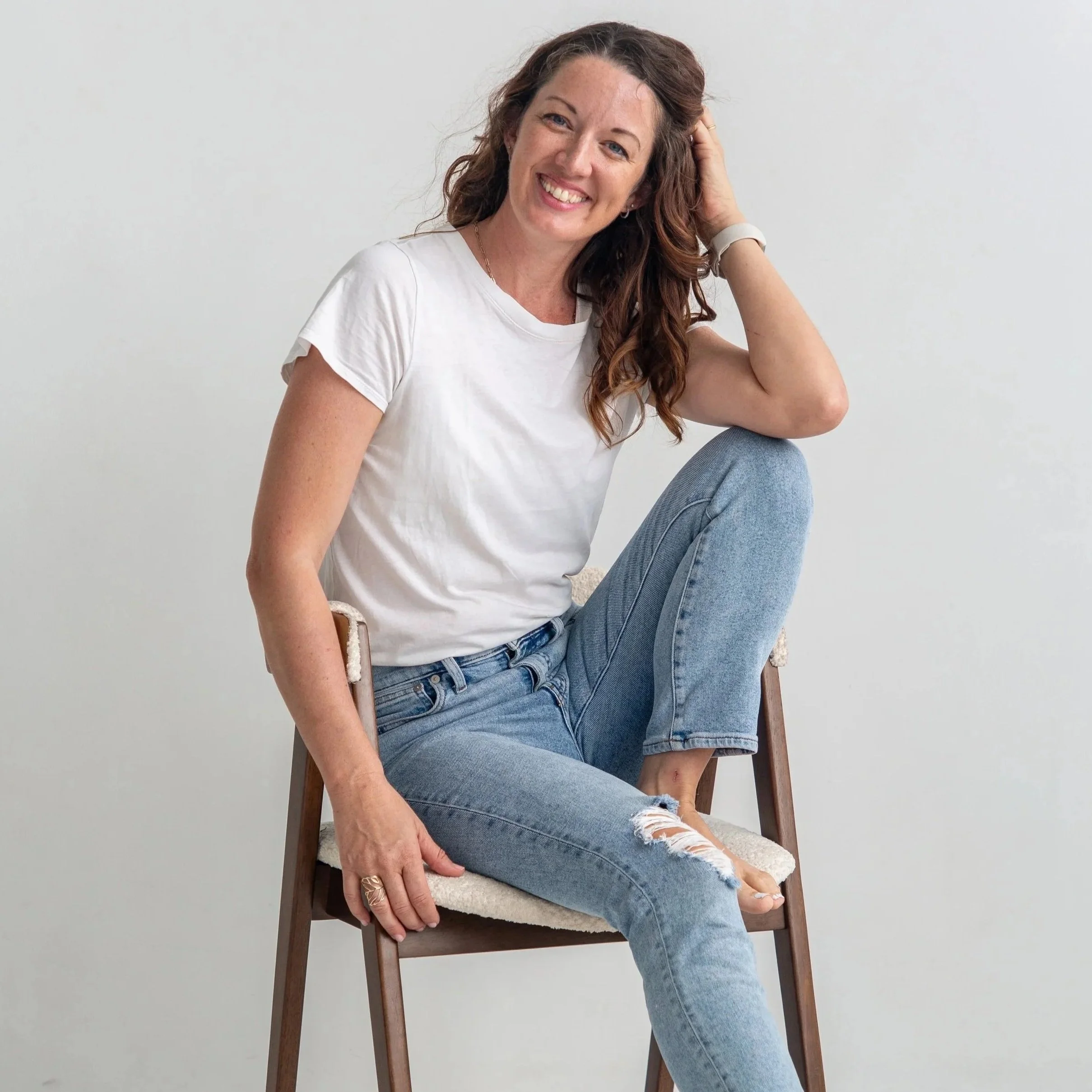 Joanie Johnson, with curly, auburn hair smiling while sitting on a wooden chair, wearing a white t-shirt and ripped jeans, with a plain light gray background.
