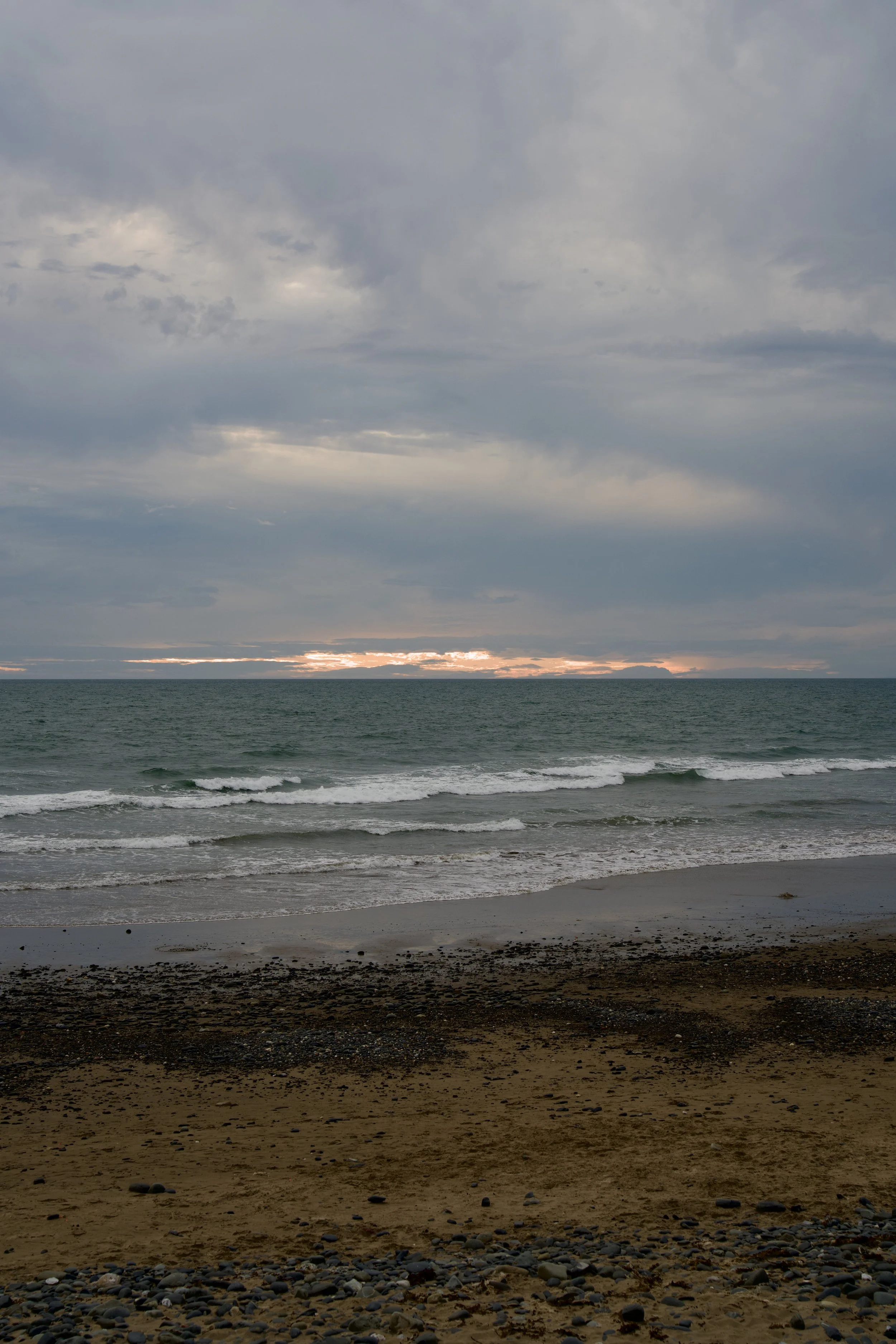 A beach scene during sunset with a cloudy sky, ocean waves, and a distant landmass on the horizon.