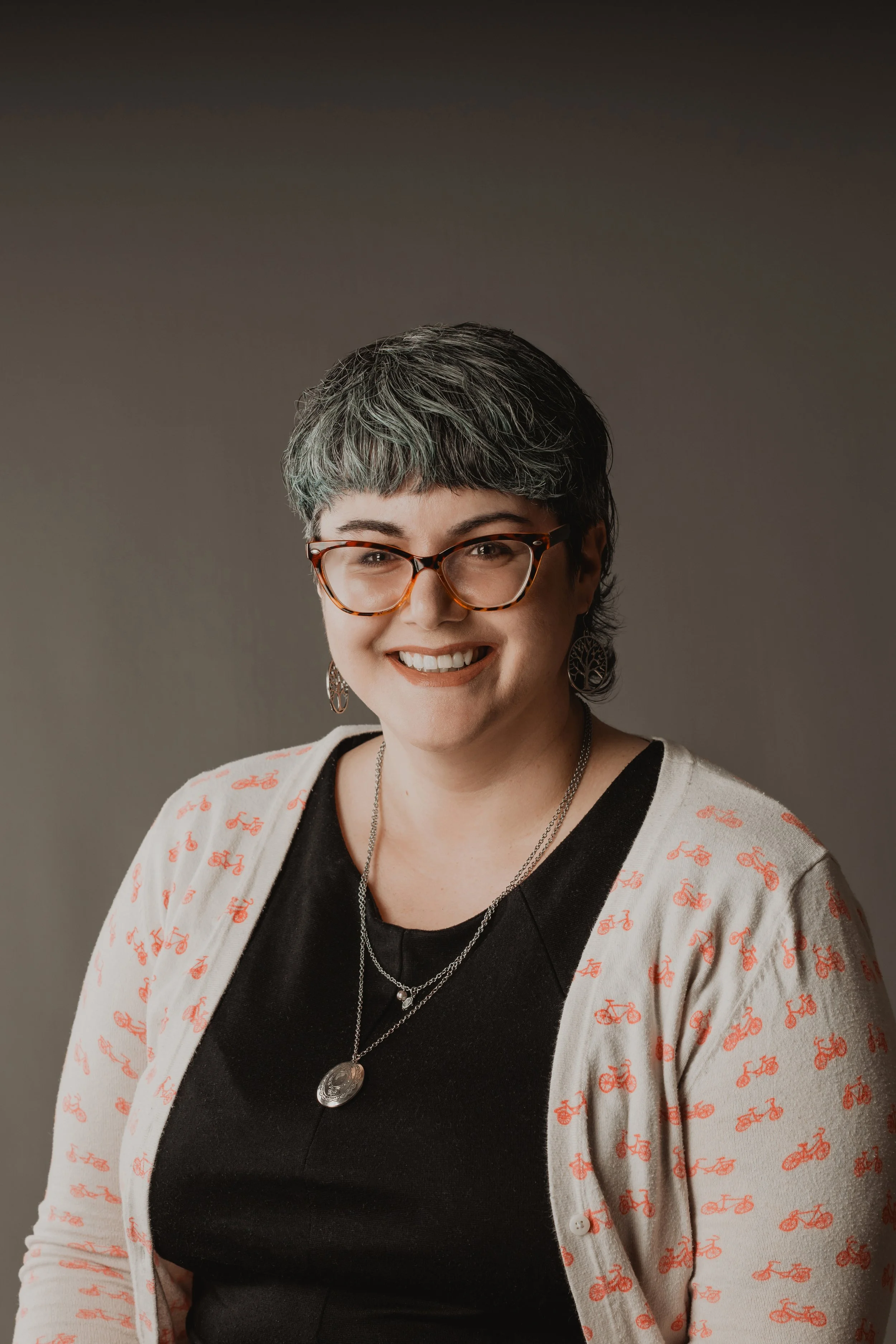 A woman with short, gray and black hair, wearing glasses, earrings, and layered necklaces, smiling at the camera. She is dressed in a black top and a white cardigan with red bicycle patterns, against a neutral gray background.