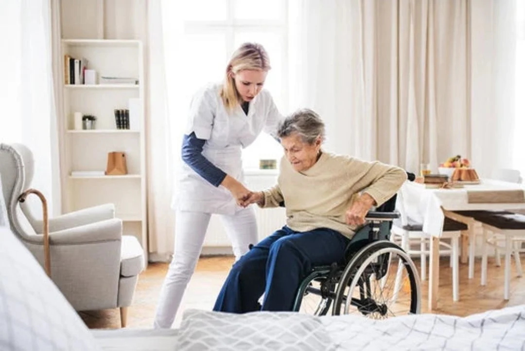 Nursing assistant helping an elderly woman in a wheelchair in a bright, home-like room.