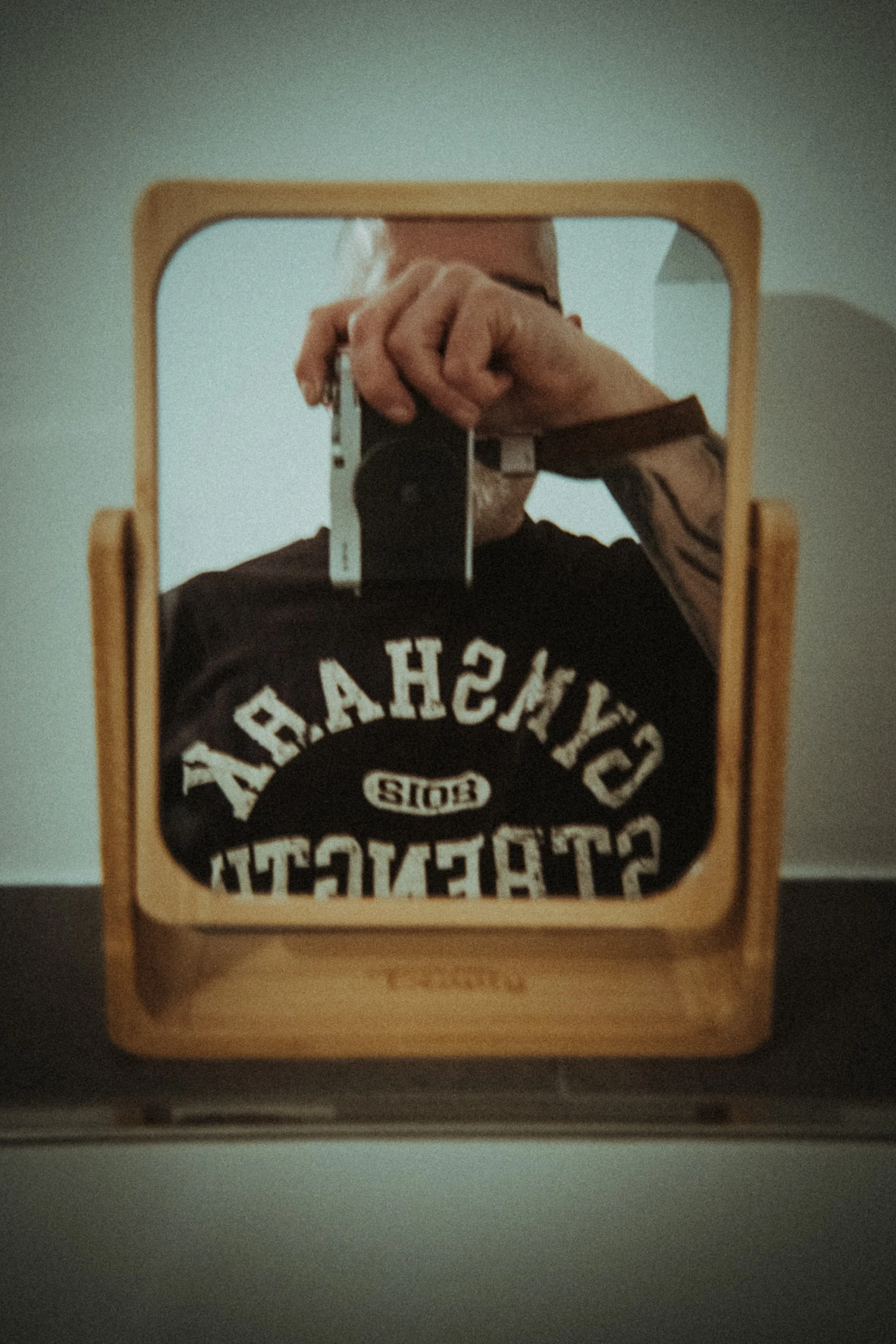 Person taking a mirror selfie using a camera, wearing a black T-shirt with white text, and a tattoo on their arm. The mirror has a wooden frame.