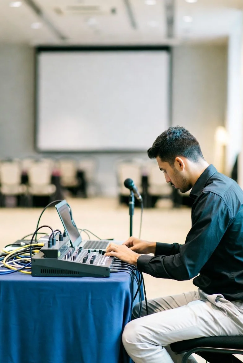 A man operating audio equipment at a table with a laptop and microphone, seated in a large room with a blank projector screen in the background.
