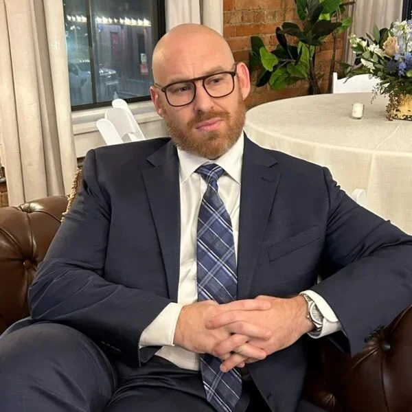 Jeff Strauss sitting on a couch wearing a suit, shaved head, brown beard, wearing glasses with black frames. Listening intently with hands and legs crossed.