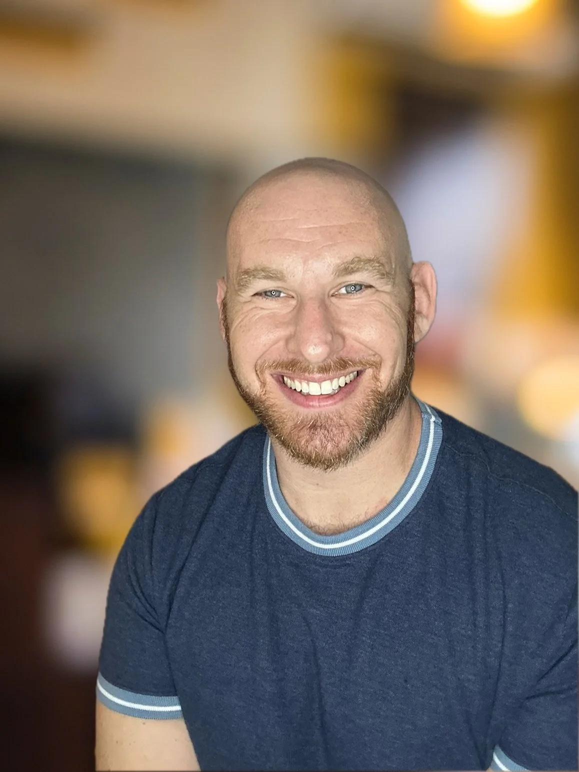 Headshot of Jeff Strauss. Smiling, shaved head, brown beard, wearing a blue knit short-sleeved shirt against a blurred background.