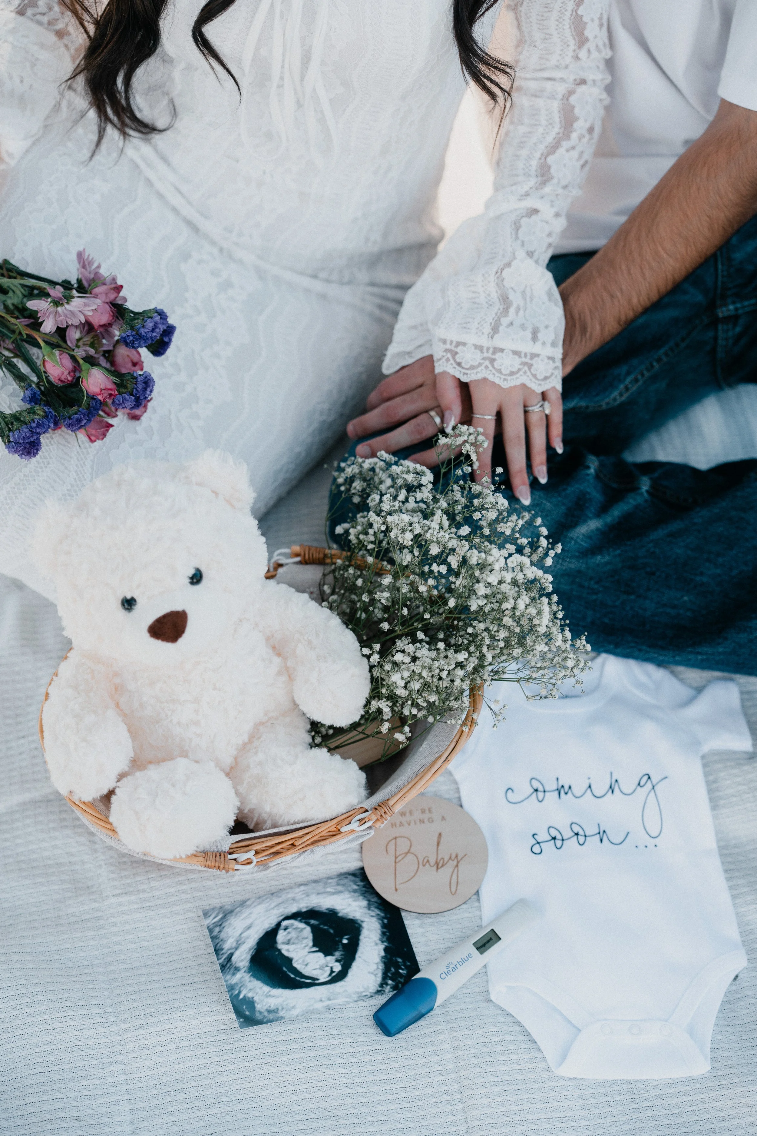 Pregnant woman and partner holding hands with visible rings, surrounded by baby items including teddy bear, flowers, ultrasound picture, 'coming soon...' onesie, and a positive pregnancy test.