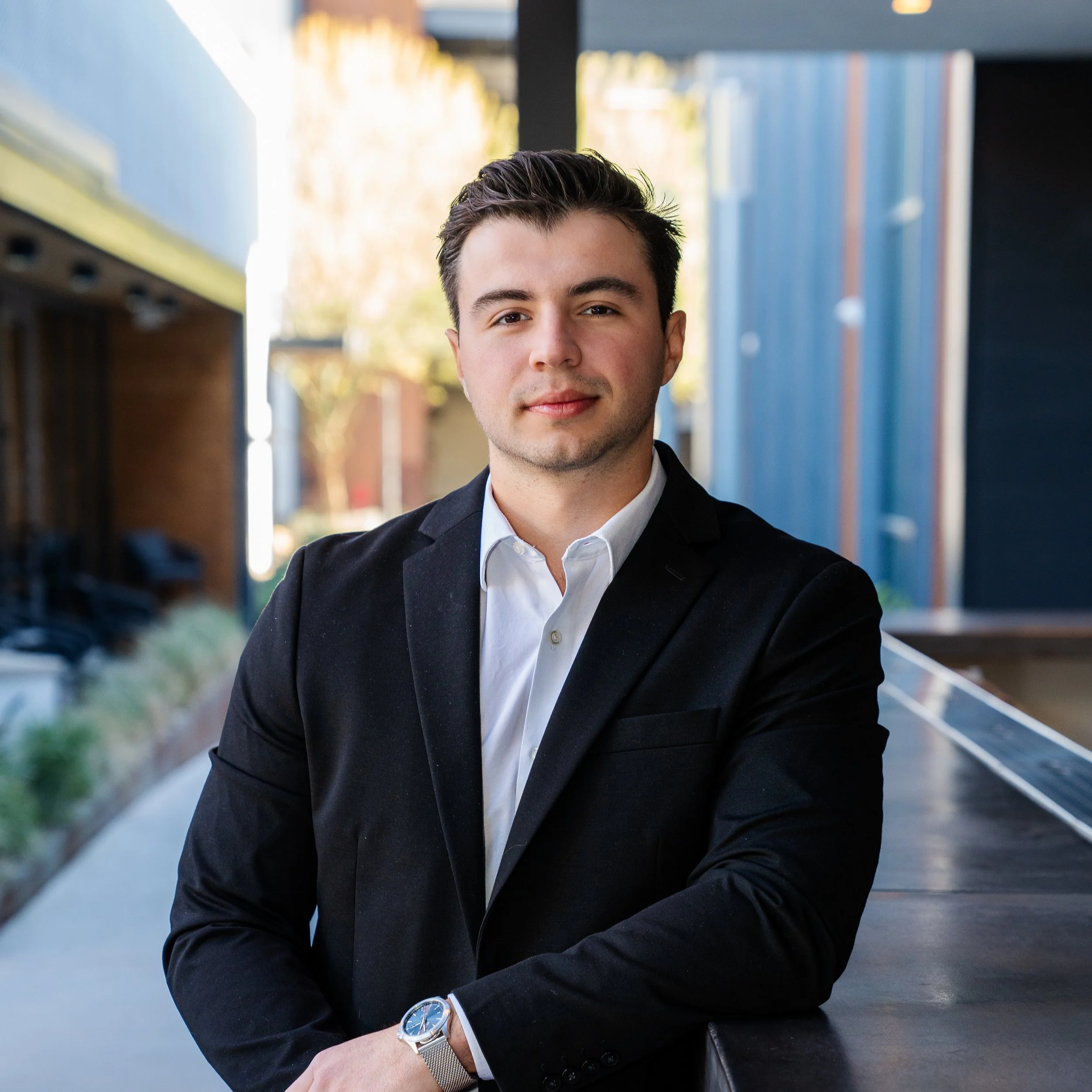 Young man with dark hair in a black suit and white shirt, standing outdoors, arms crossed, with a modern urban background.