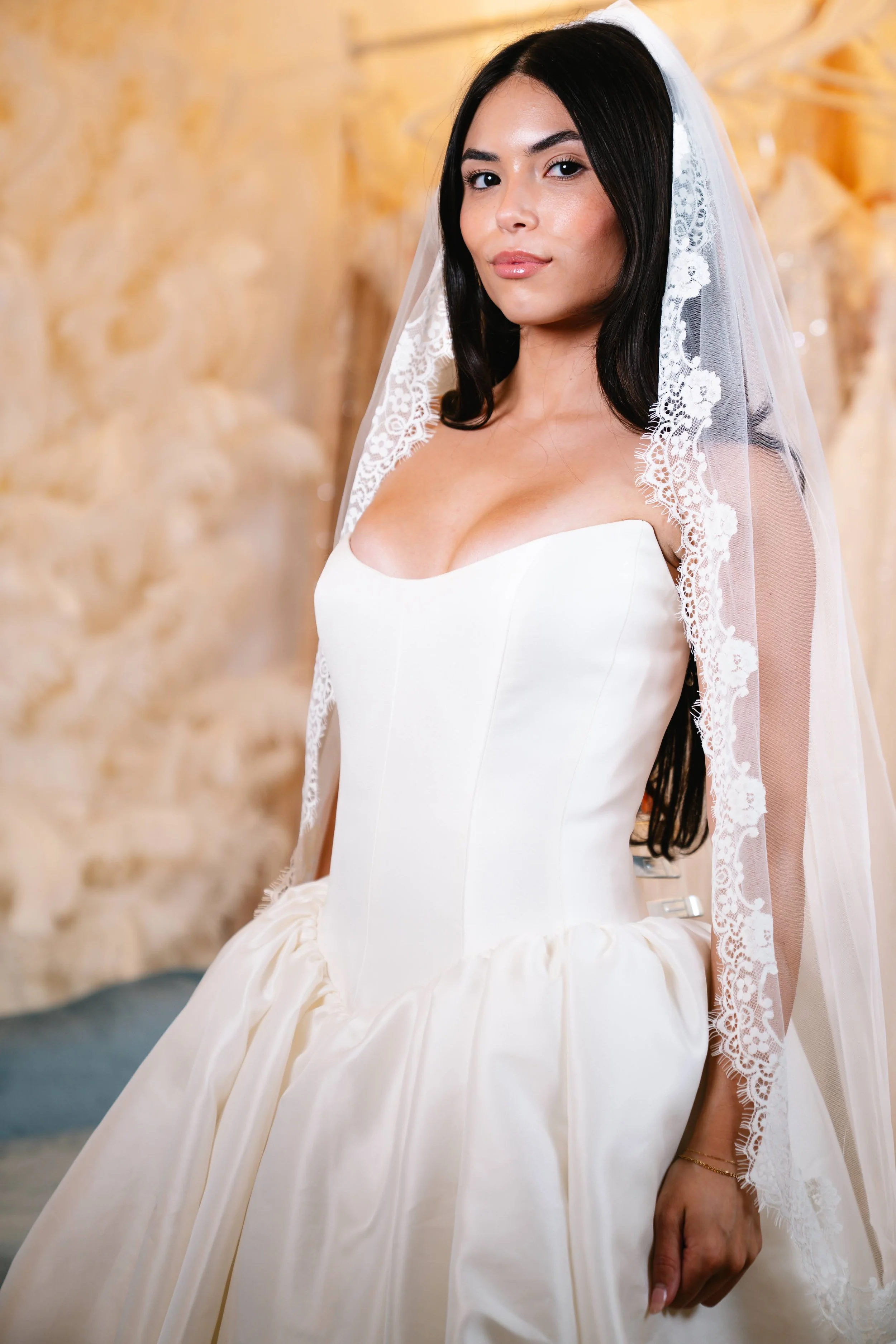 A woman in a white wedding dress and veil standing against a beige textured wall.