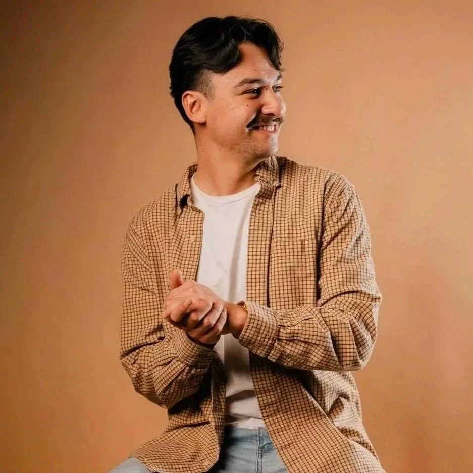 A young man with dark hair, a mustache, and a beard, smiling and looking to his left. He's wearing a beige checkered shirt over a white T-shirt and appears to be clapping or rubbing his hands together. The background is a solid tan color.