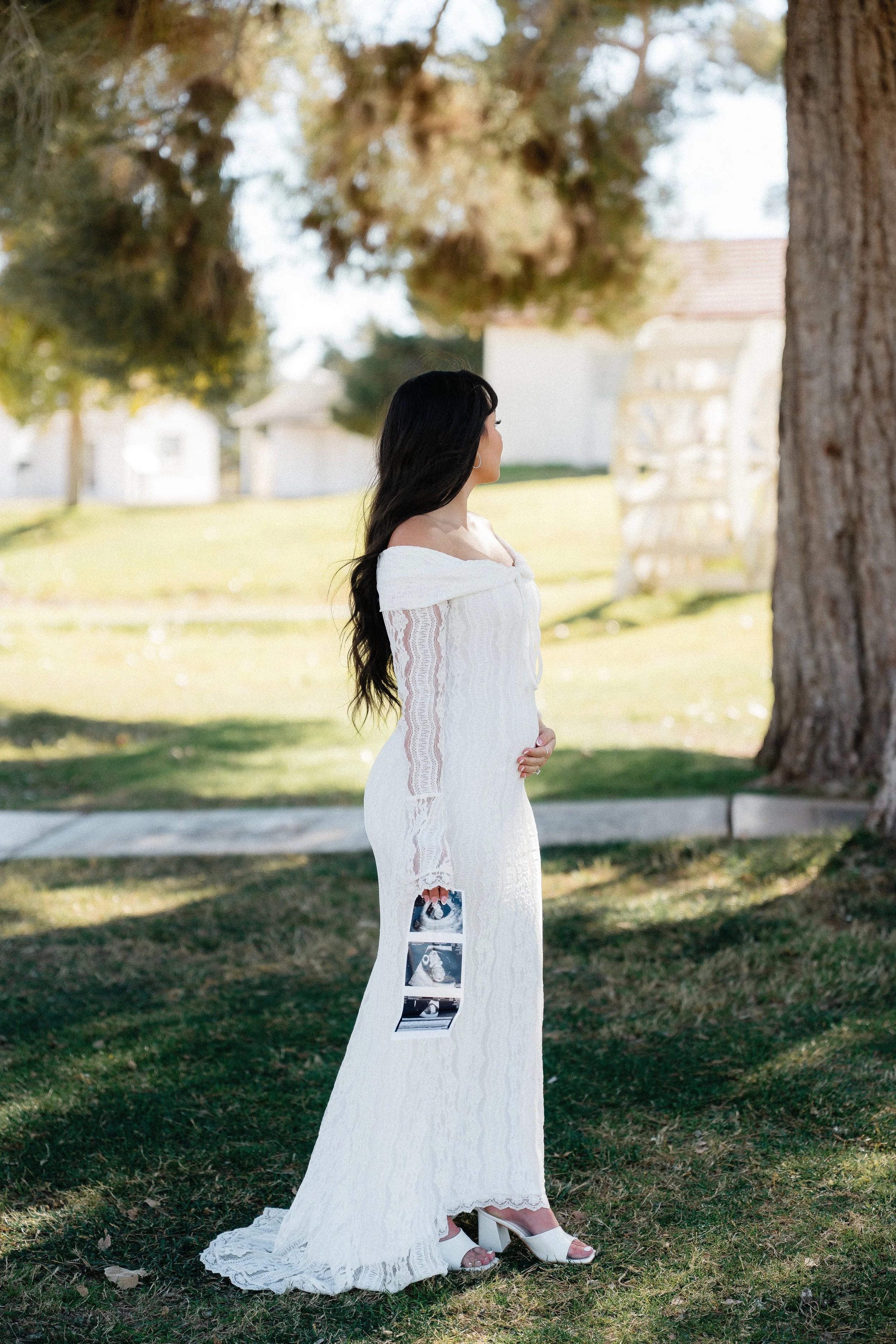 A woman dressed in a white lace dress standing outdoors near a large tree, holding ultrasound pictures.