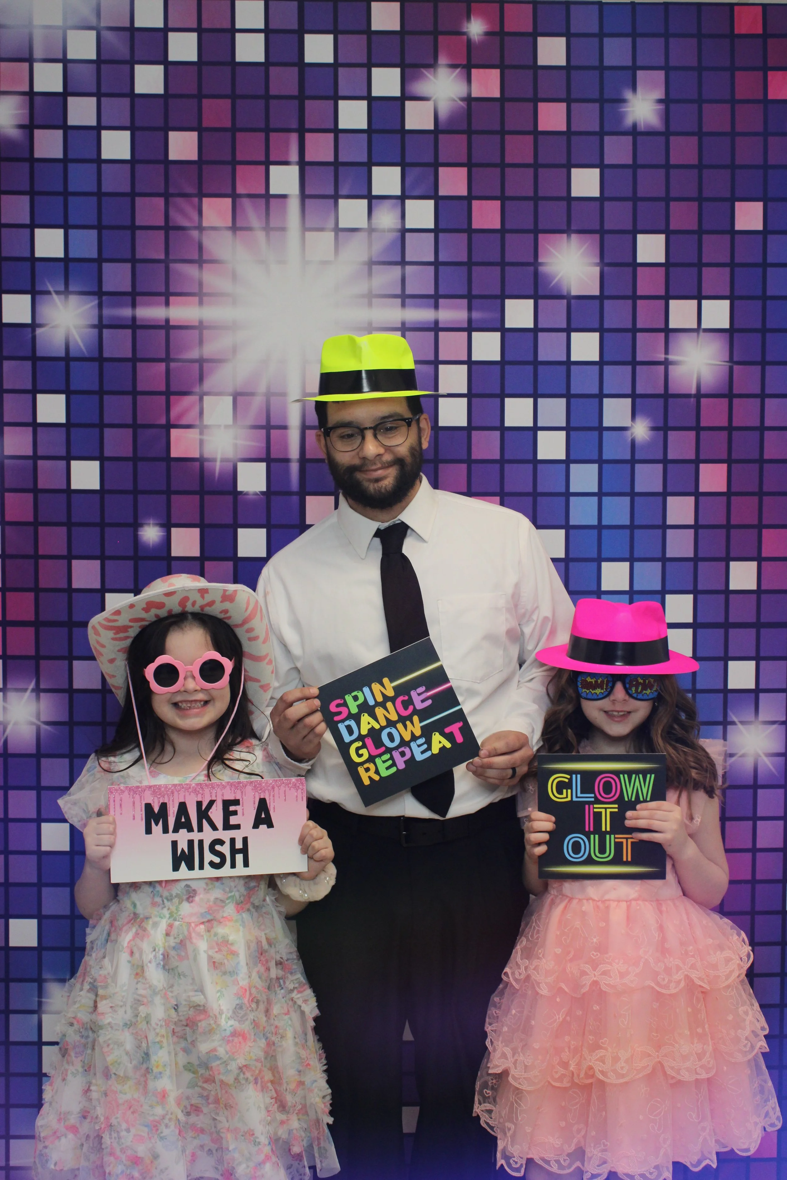 Three people at a colorful, shiny, disco-themed photo booth: a man in a white shirt and black tie with neon yellow hat, and two girls in frilly dresses and oversized sunglasses, holding signs with dance-themed messages.