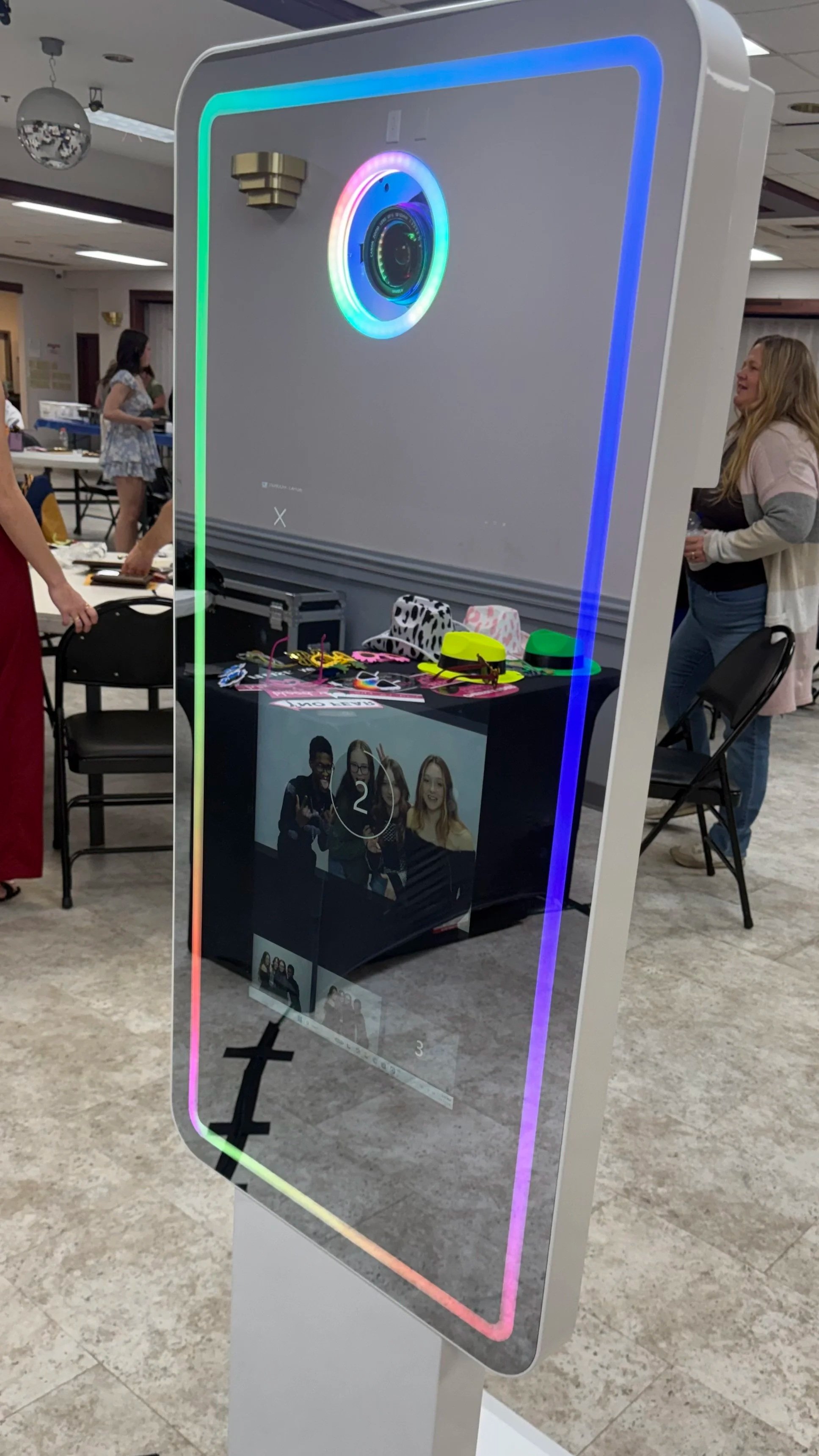 A photo booth with a digital screen displaying a photo of four women making peace signs. The booth has colorful rainbow lights around the camera and a bright neon border. In the background, people are standing and chatting inside a room.