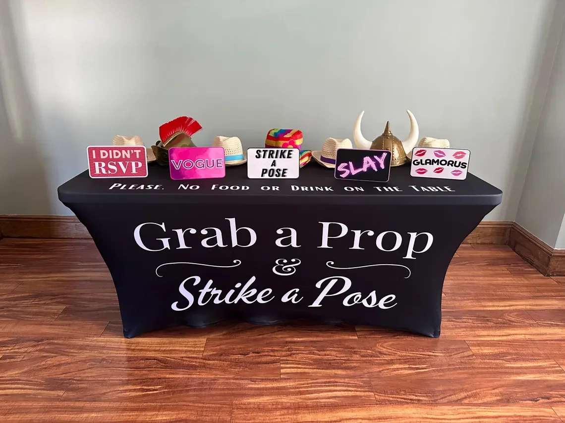 Table with props and signs for a photo booth, including hats, a horn, signs with phrases like "I DIDN'T RSVP," "VOGUE," "STRIKE A POSE," "SLAY," and "GLAMOROUS," with a black tablecloth displaying the phrase "Grab a Prop & Strike a Pose."