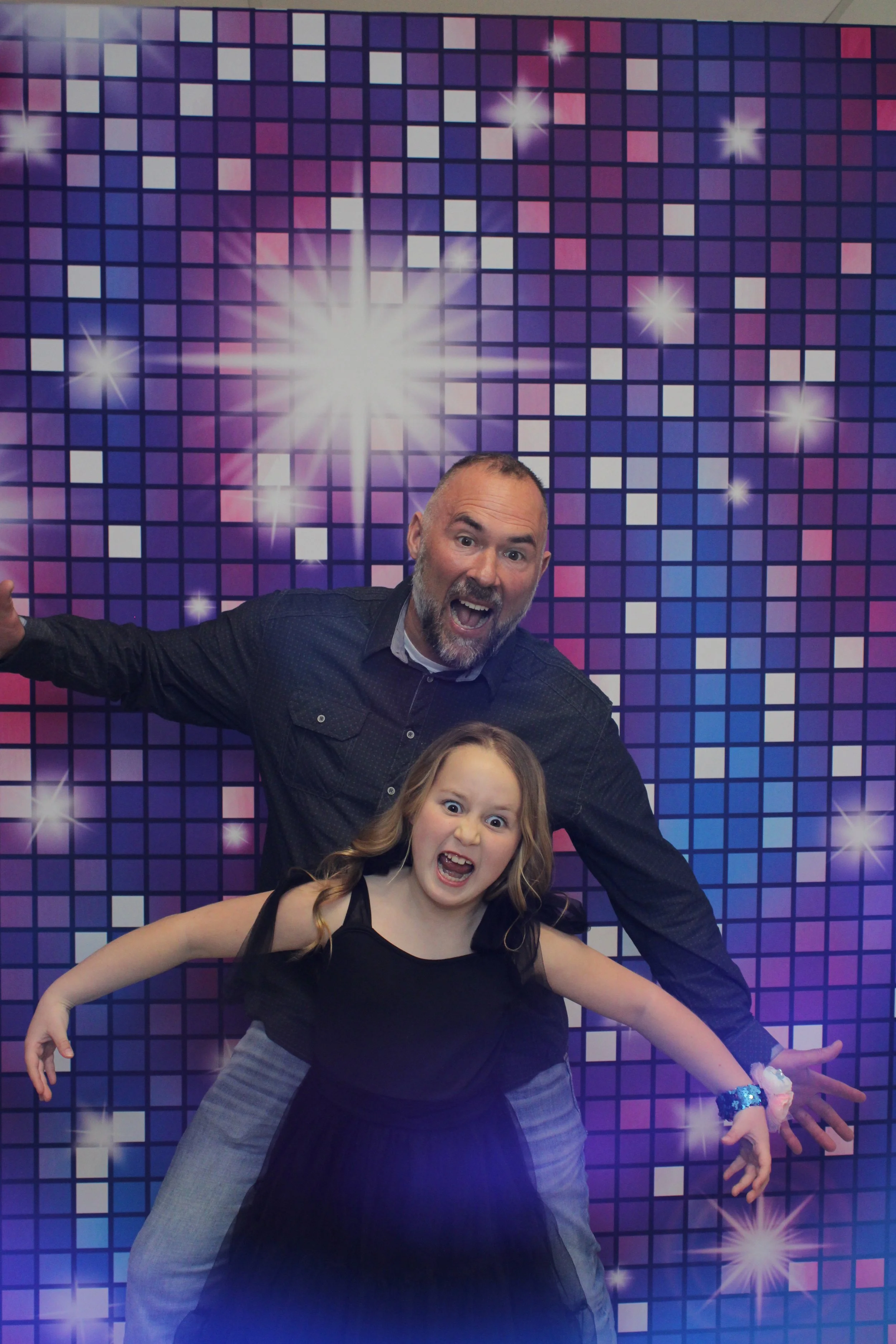 Man and girl making silly faces and pose in front of a disco-themed backdrop with purple and blue shading, glowing stars, and a large illuminated star at the center. this was took at a daddy daughter dance in norwalk ohio.