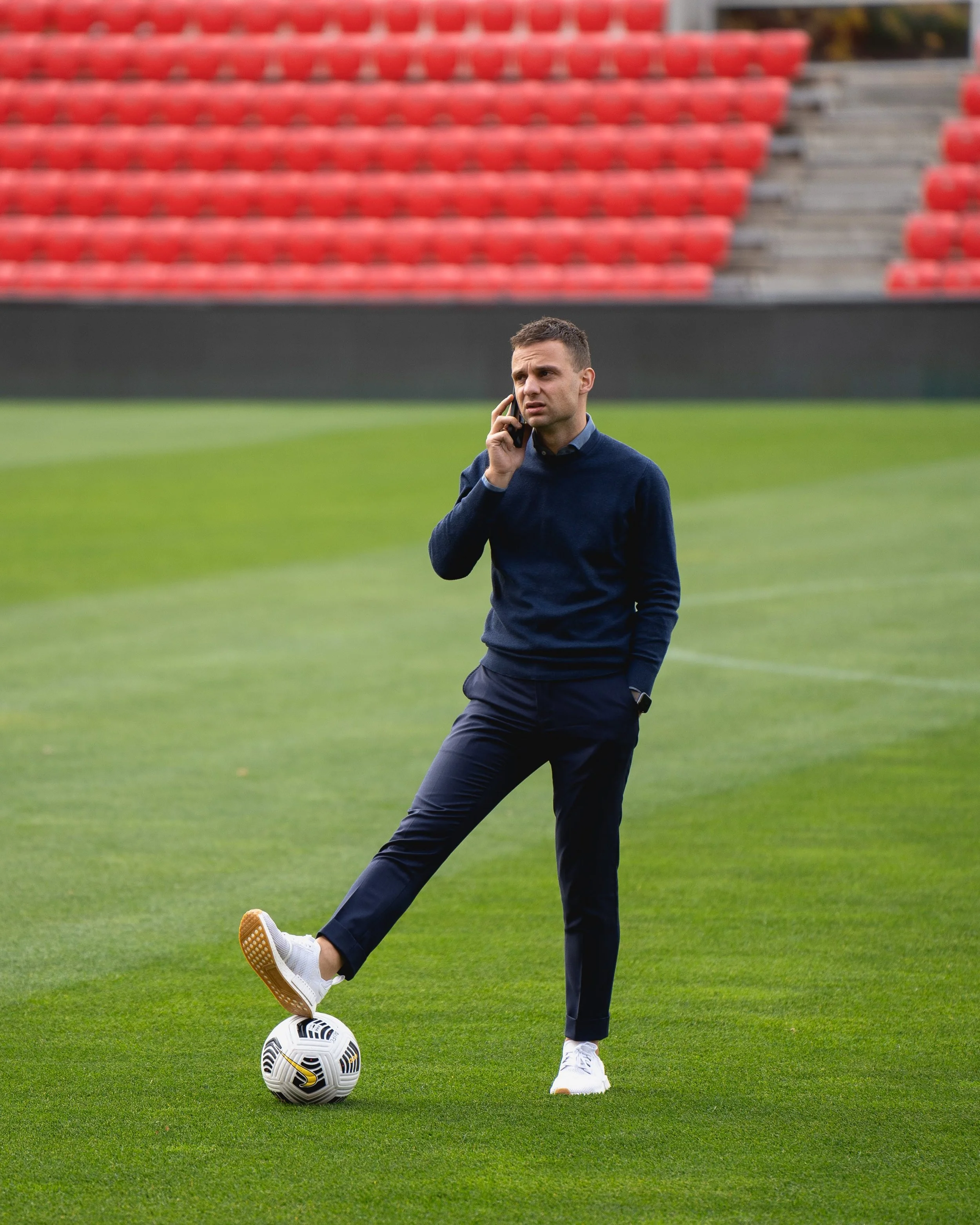 A man dressed in a dark blue sweater, pants, and white sneakers is standing on a soccer field, with one foot resting on a soccer ball. He is holding a phone to his ear, appearing to be engaged in a conversation. The background features rows of red stadium seats.