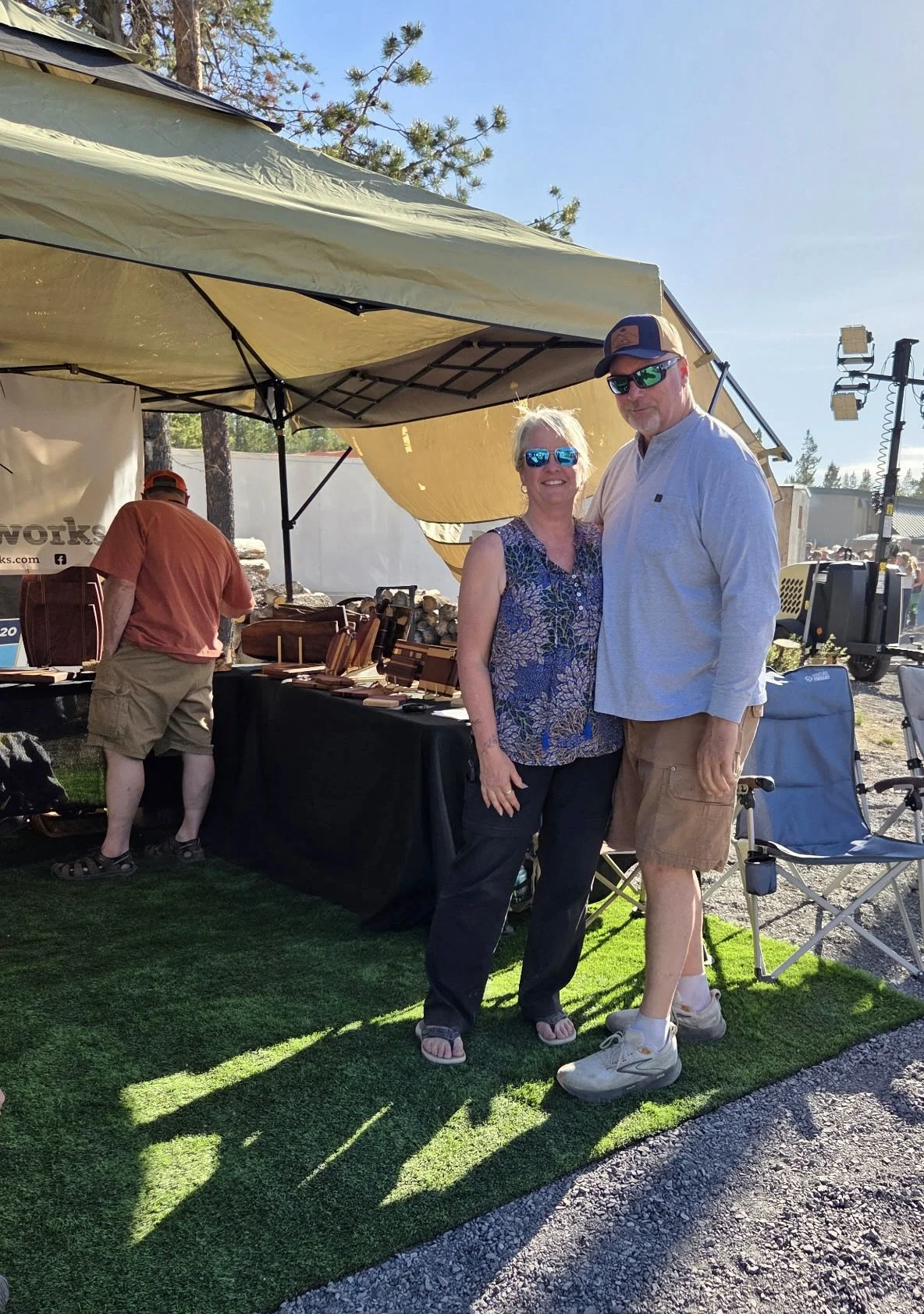 The owners of Two Rivers North Woodworks stand together outdoors at a market next to their vendor booth with handmade wooden products.