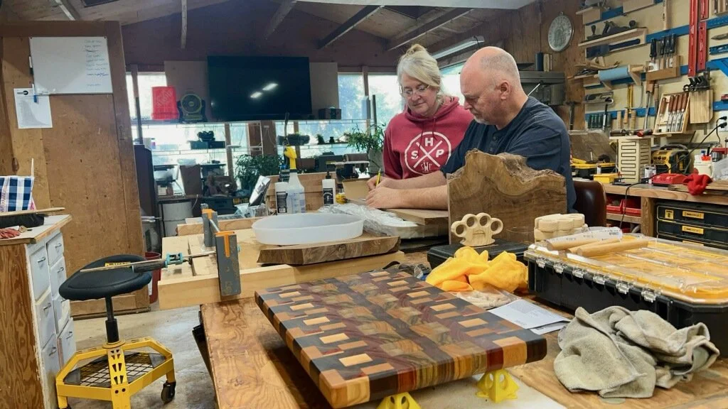Two individuals working in a woodworking shop surrounded by wood pieces, tools, and supplies.