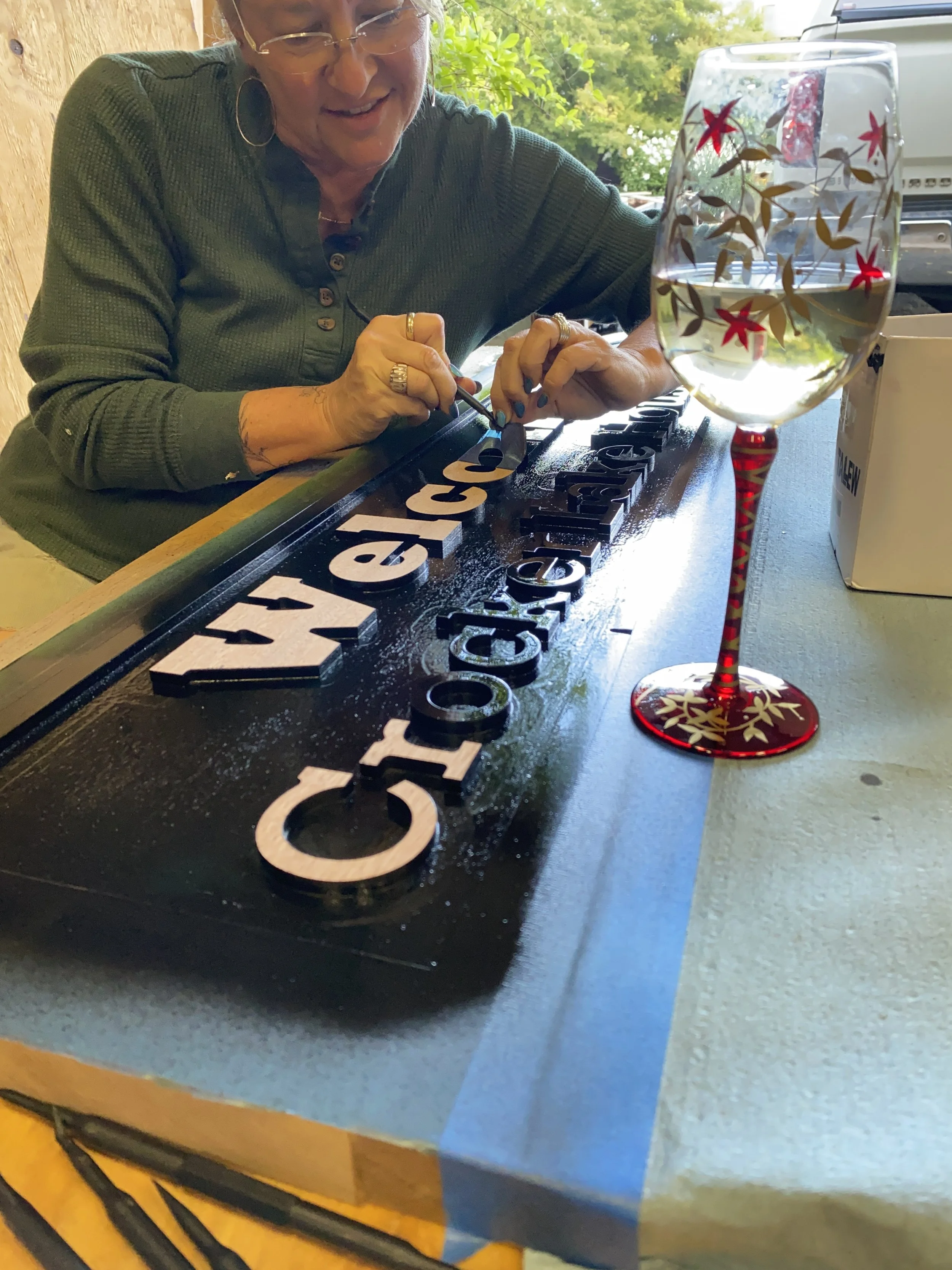 A woman decorating a handcrafted sign wooden letters at a table outside with a glass of water in the foreground.