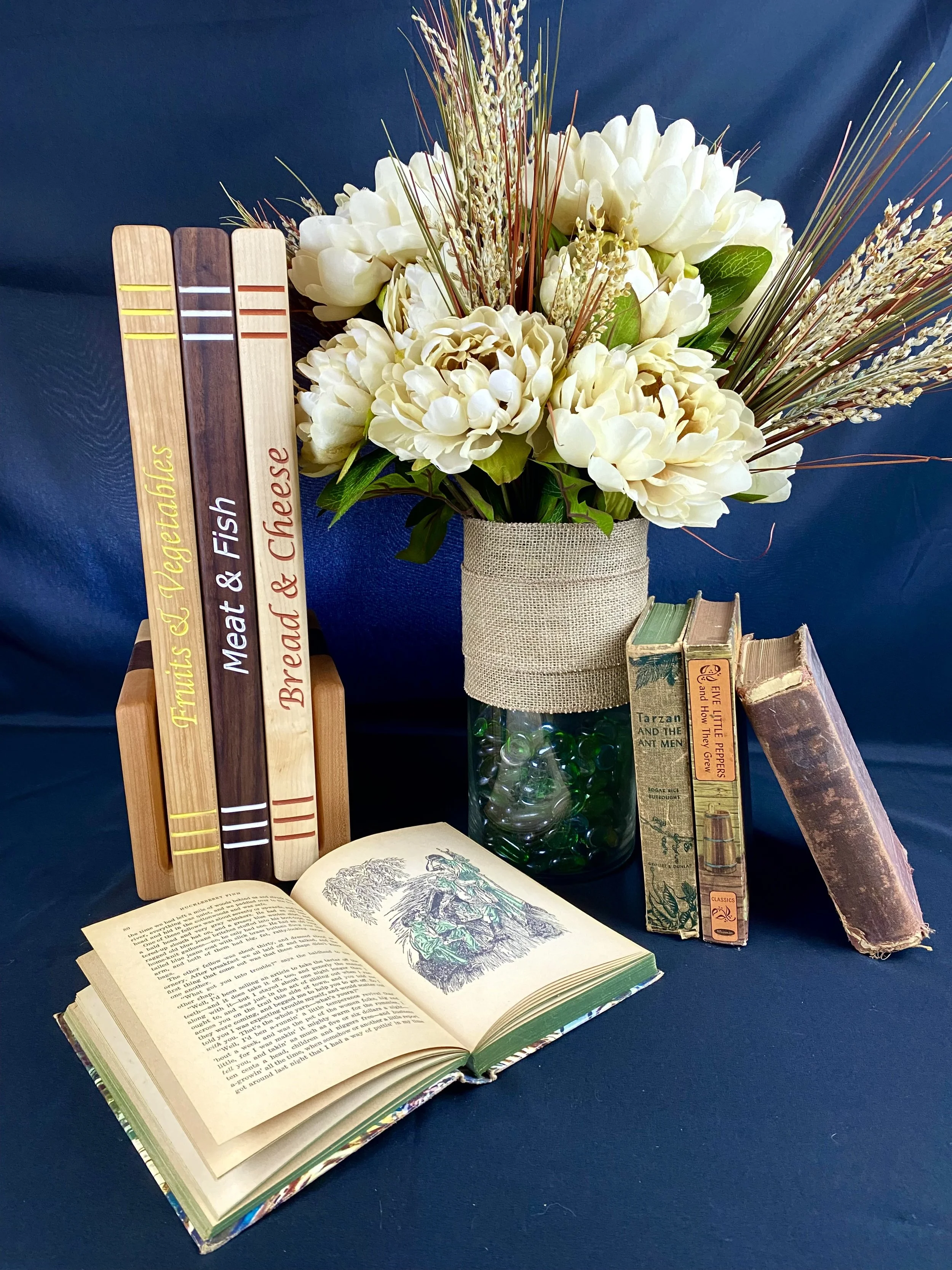 A floral arrangement with white flowers and tall grasses in a burlap-covered vase, an open book with illustrations and text, several hardcover books stacked upright, and wooden book-like signs with book titles, all on a blue background.