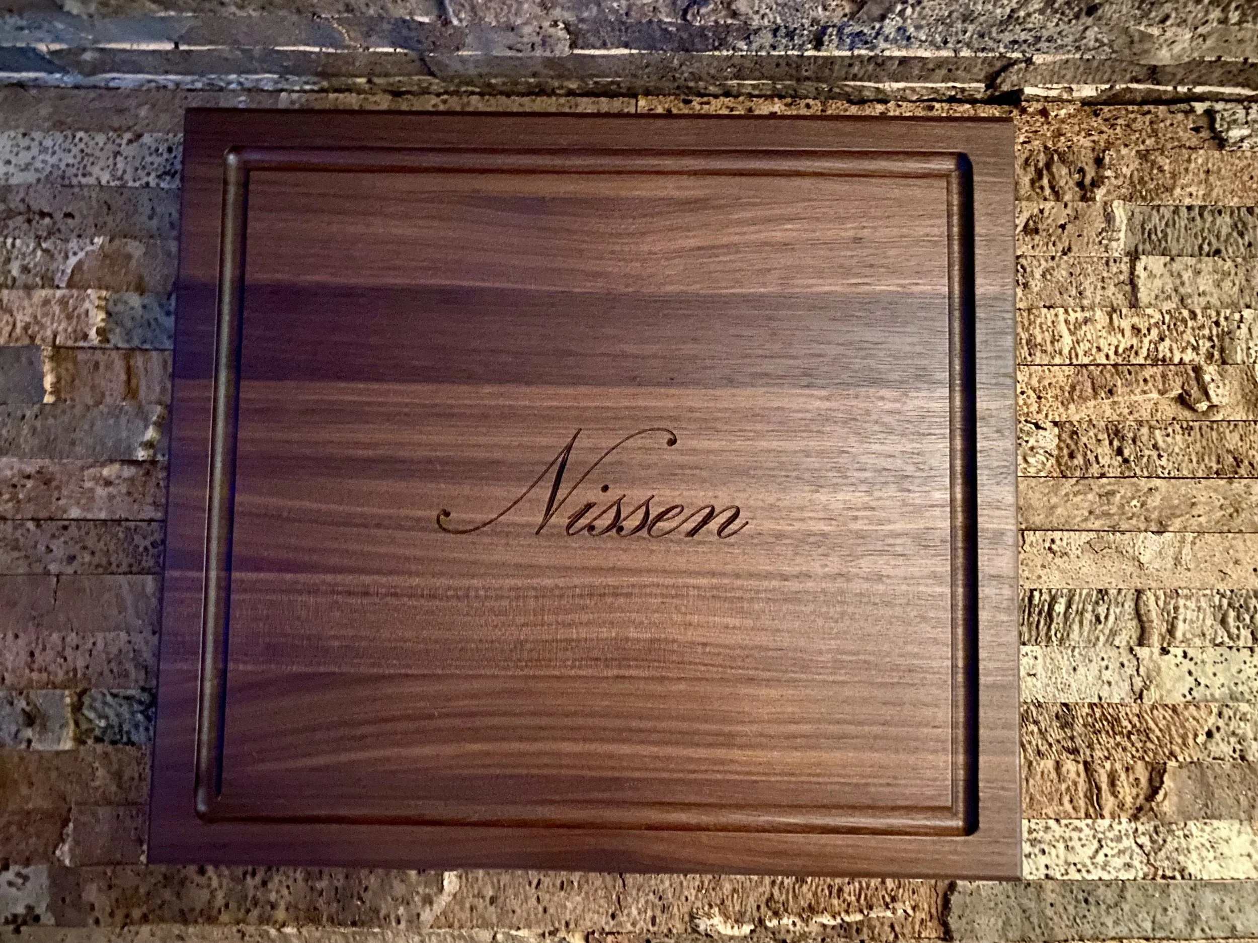 A handmade wooden sign with the word "Nissen" written in elegant script, placed against a textured stone wall background.
