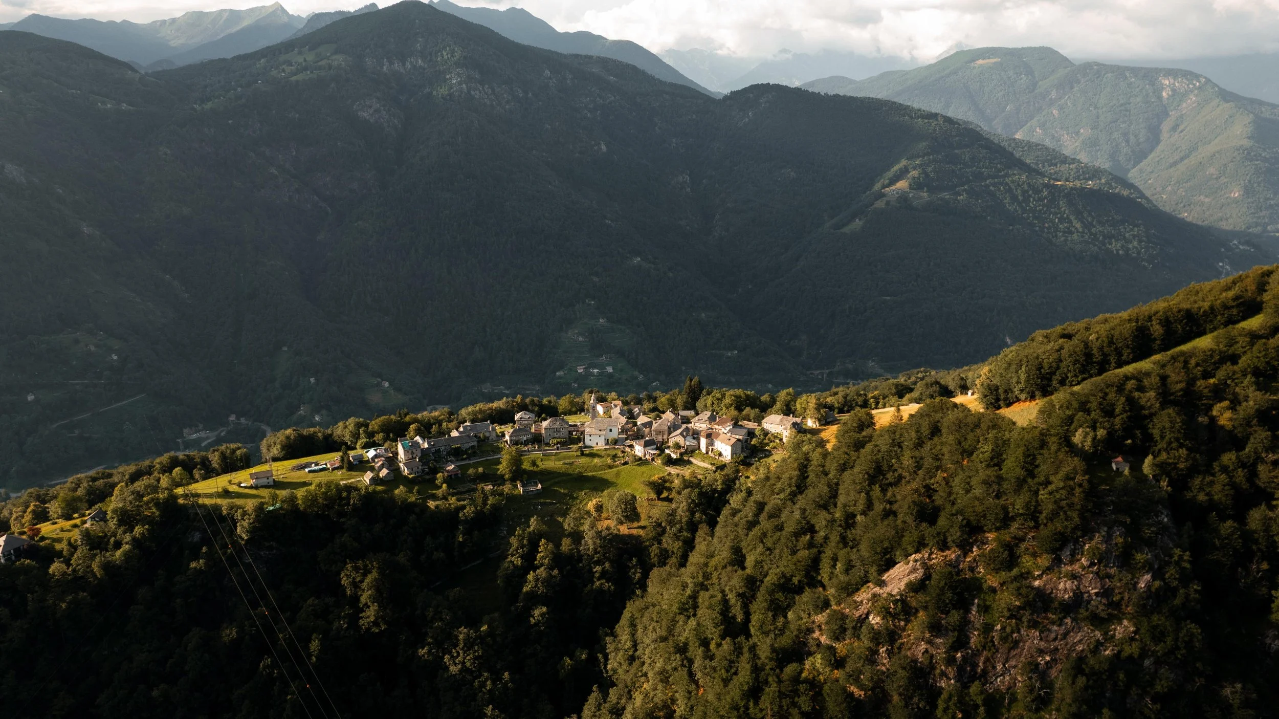 Dorf auf bewaldeter Hügellandschaft im Gebirge, umgeben von Bergen und Wäldern, bei Sonnenuntergang.