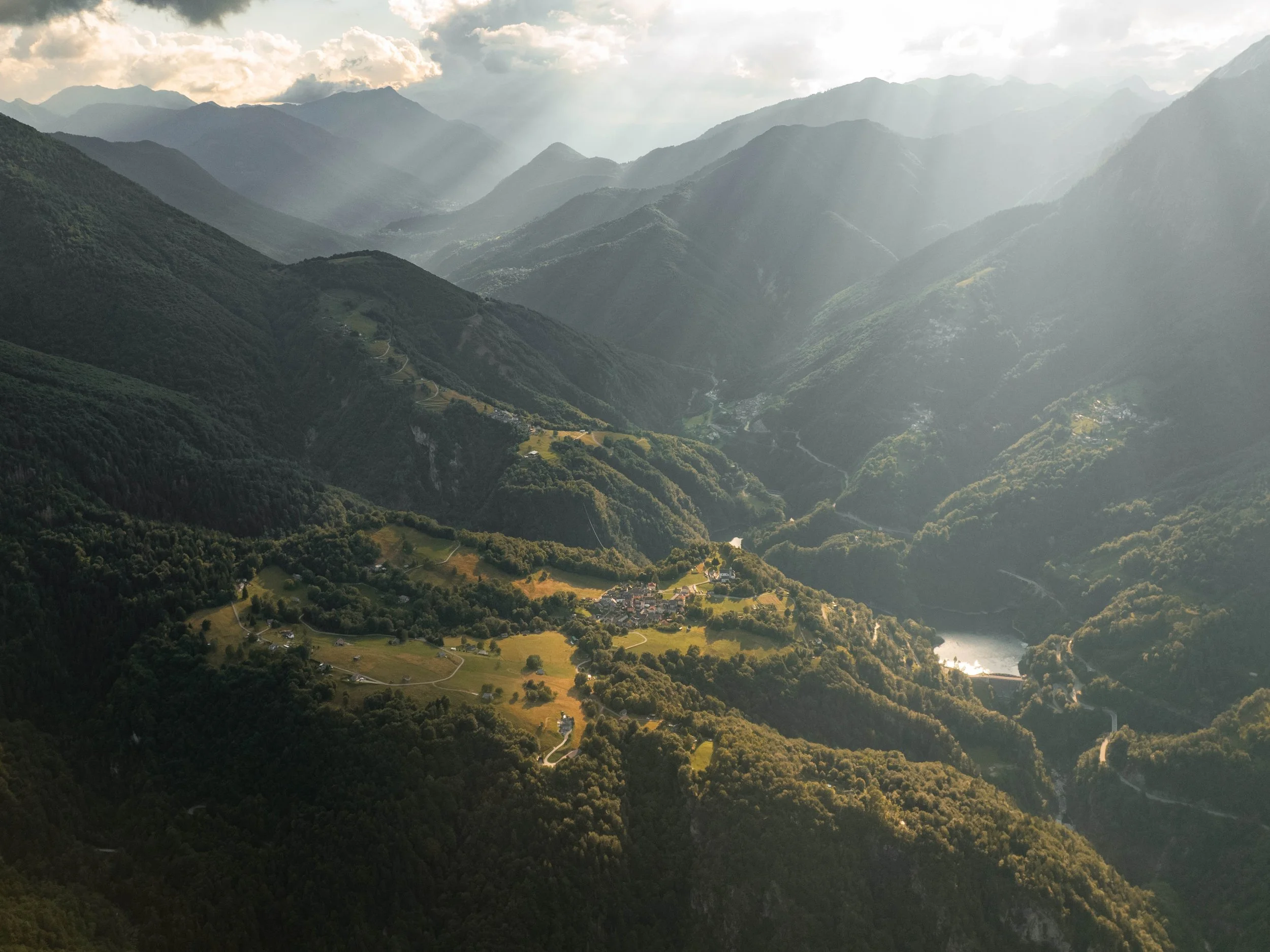 Landschaft in den Bergen mit Tal, Fluss, Häusern und bewaldeten Hügeln bei Sonnenlicht.