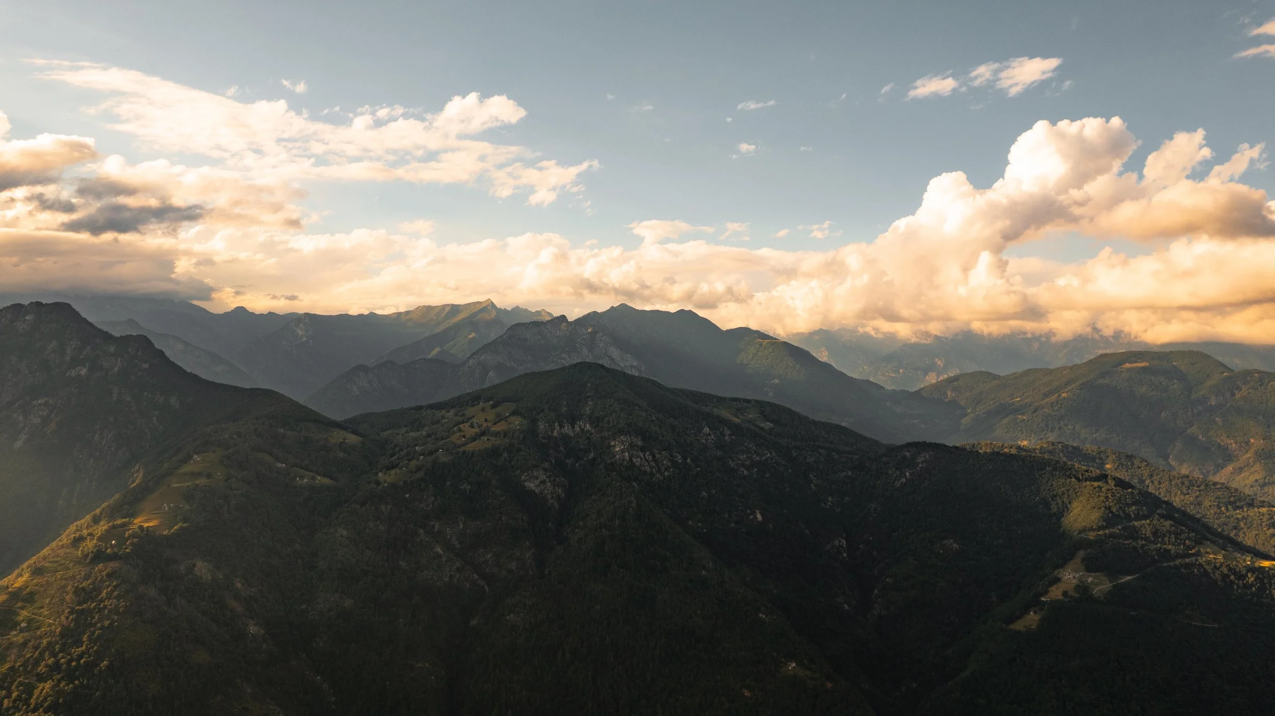 Berglandschaft mit bewaldeten Hängen, mehreren Gipfeln im Hintergrund, bewölktem Himmel bei Sonnenuntergang.