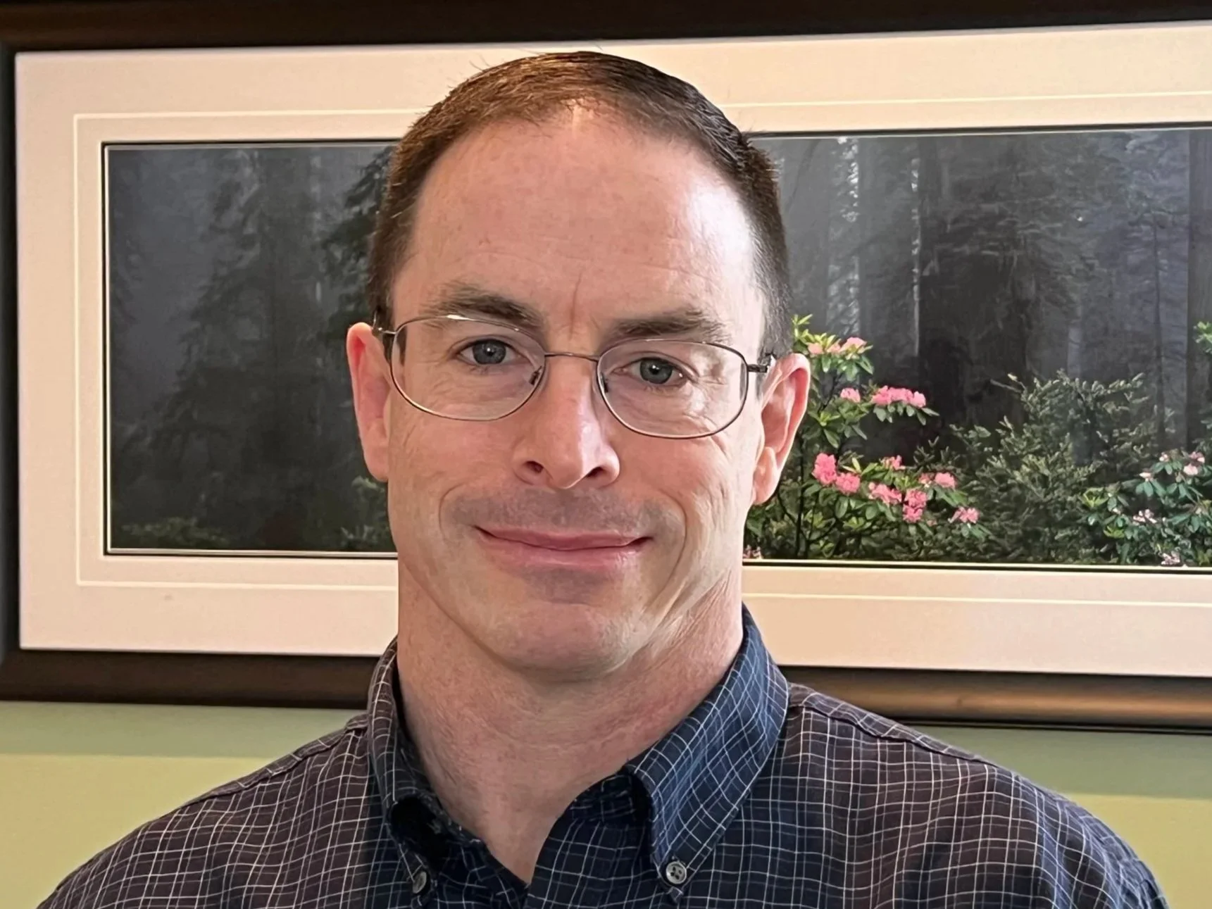 A man with short brown hair, glasses, and a checkered shirt smiling in front of a framed nature photograph with pink flowers and trees.