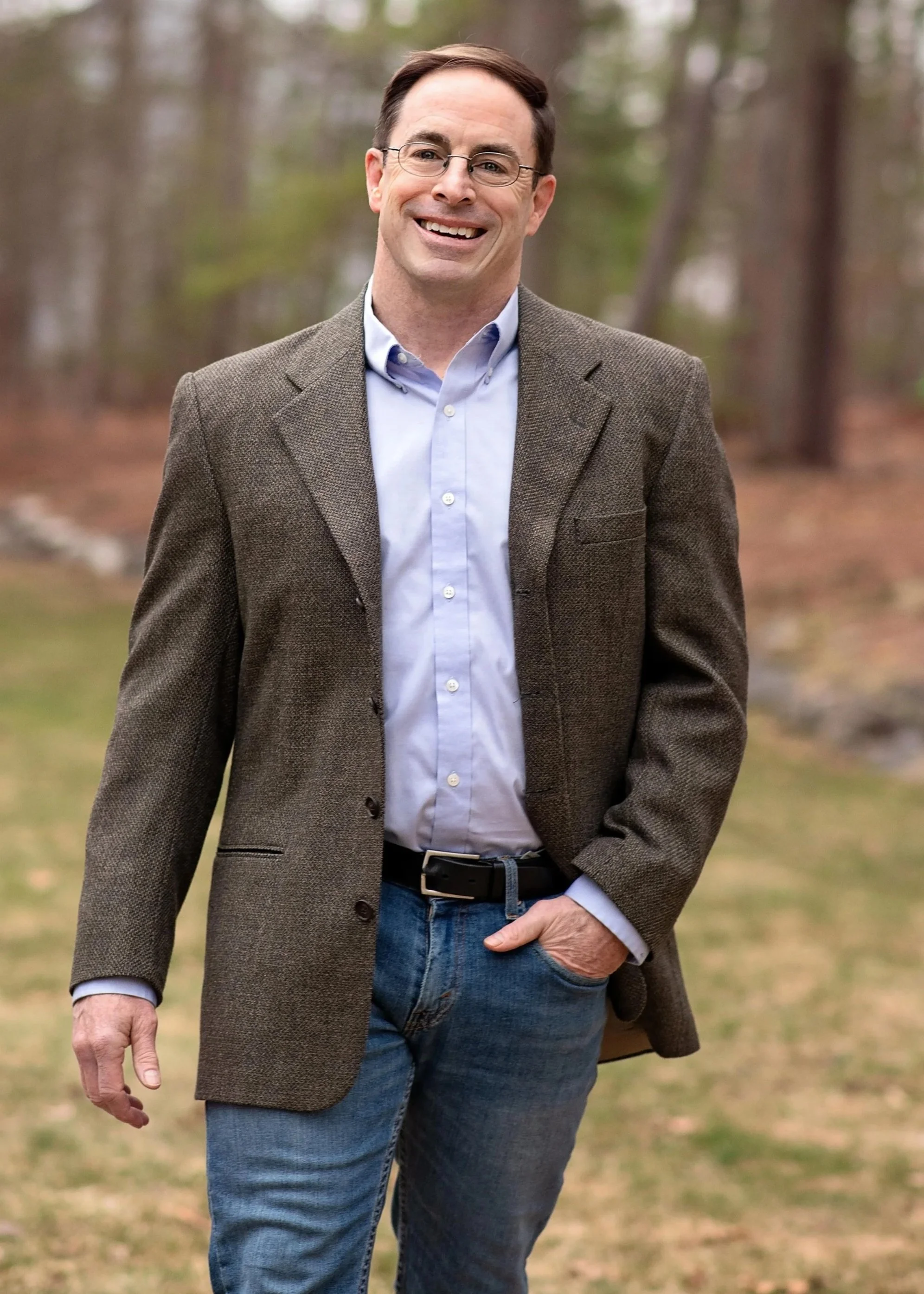 A smiling man wearing glasses, a light blue button-up shirt, a brown blazer, and blue jeans, standing outdoors in a wooded area.
