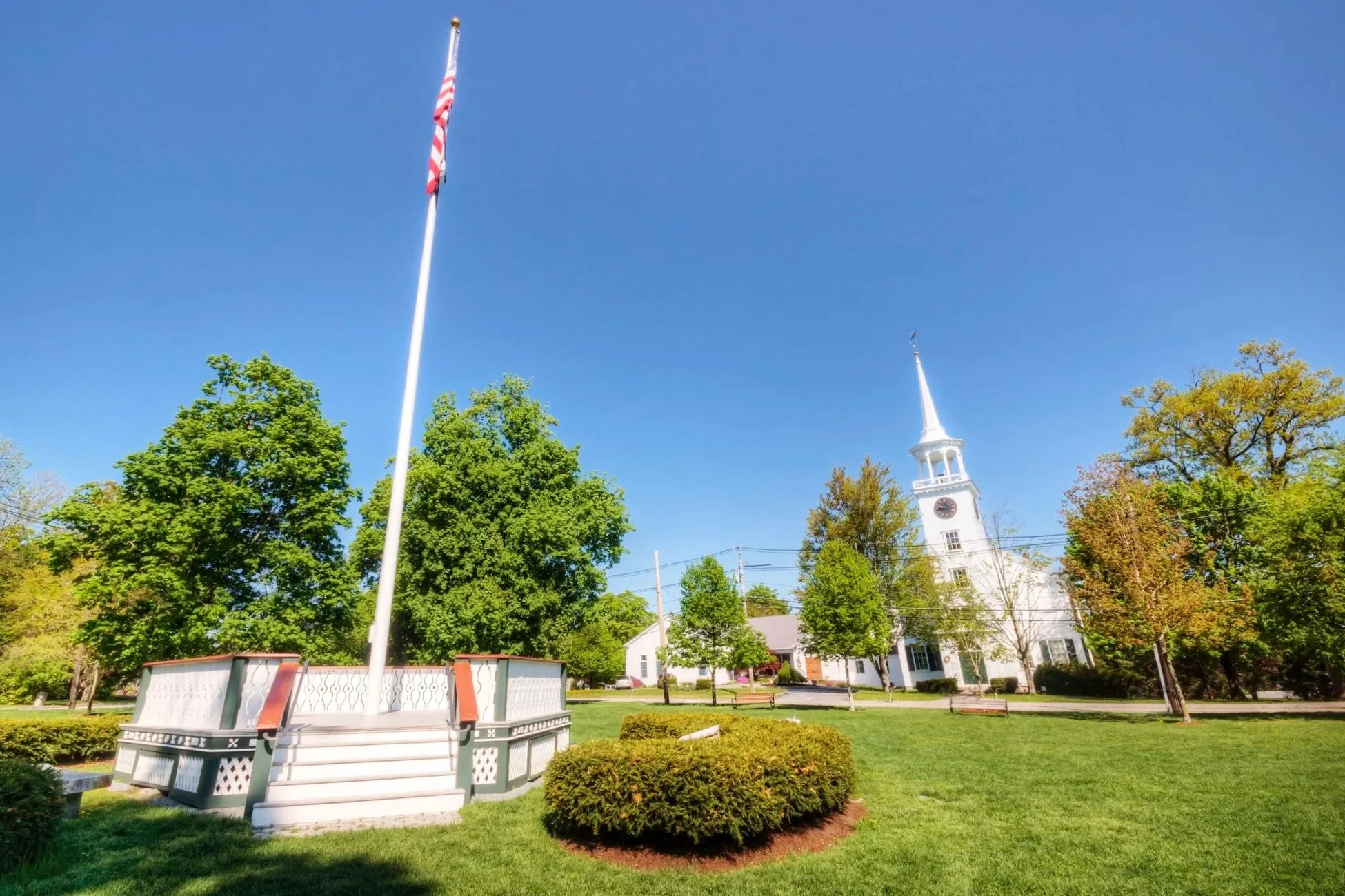 A white church with a tall steeple and a clock, surrounded by green trees and grass, under a bright blue sky. An American flag on a flagpole is in the foreground.