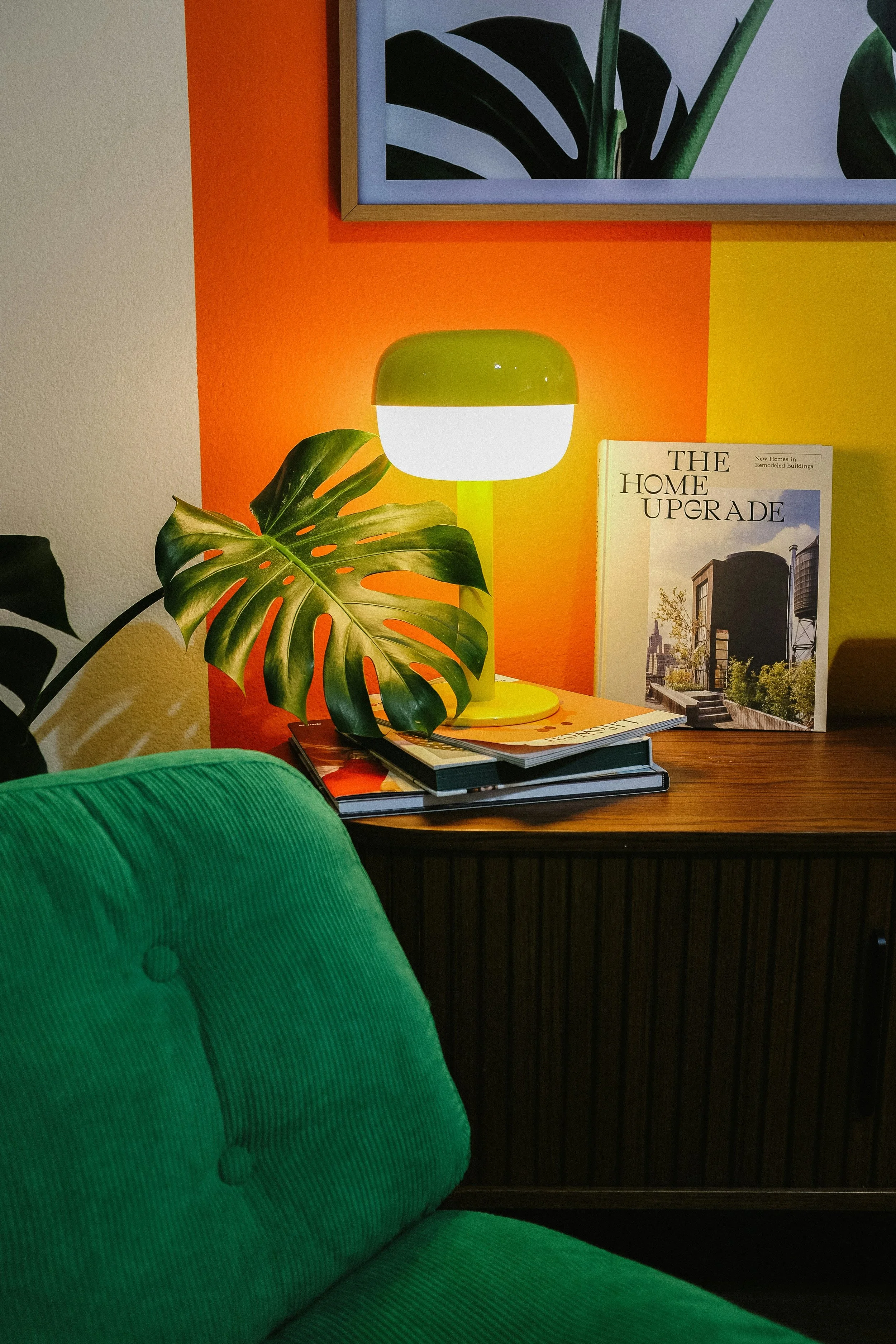 Kuva kotitoimiston pöydästä, jossa on vihreä lamppu, kirja 'The Home Upgrade', pino lehtiä ja suuri monstera-kasvin lehti, taustalla oranssi ja keltainen seinä.